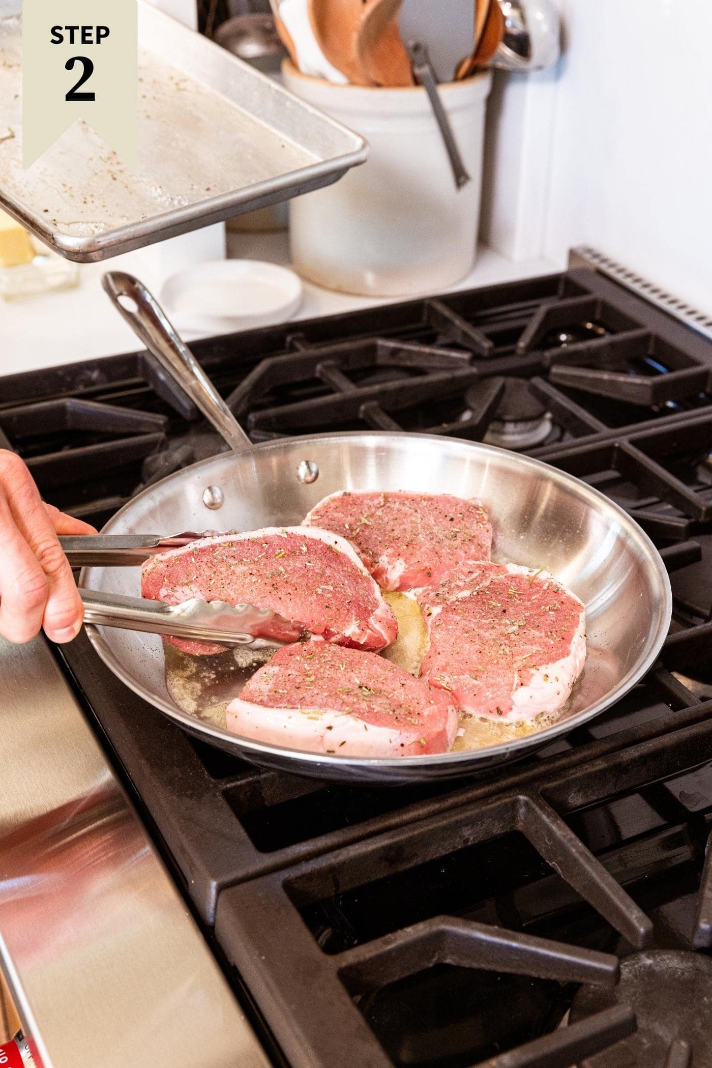Hand holding tongs browning four pork chops in metal skillet on stovetop.