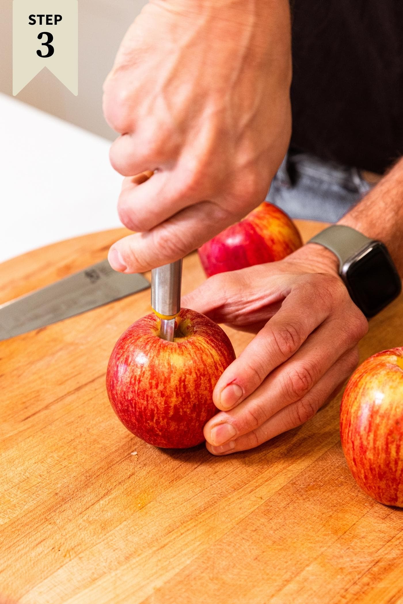 Two hands holding an apple using tool to core out the center of the apple with more apples sitting around waiting as well.