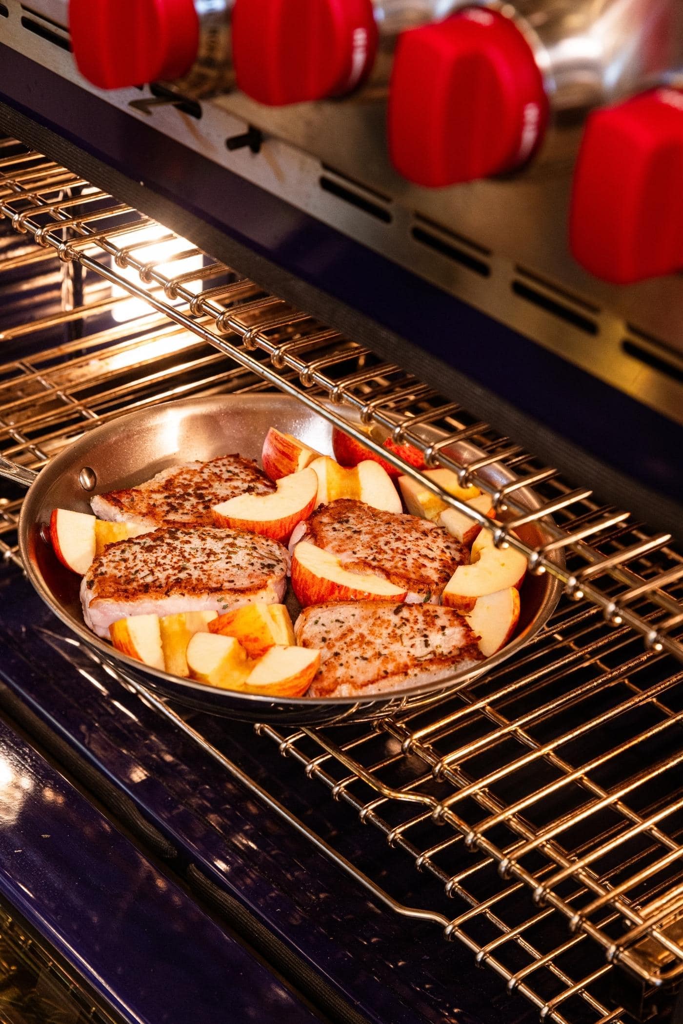 Metal skillet going into a rack in an oven with browned pork chops and raw apples inside.