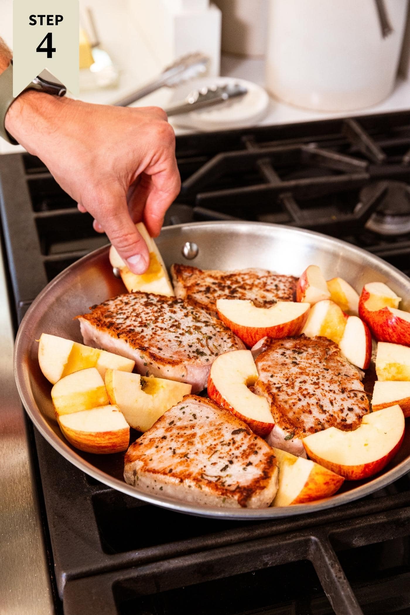 Hand placing apple slices around browned pork chops in metal skillet on stovetop.