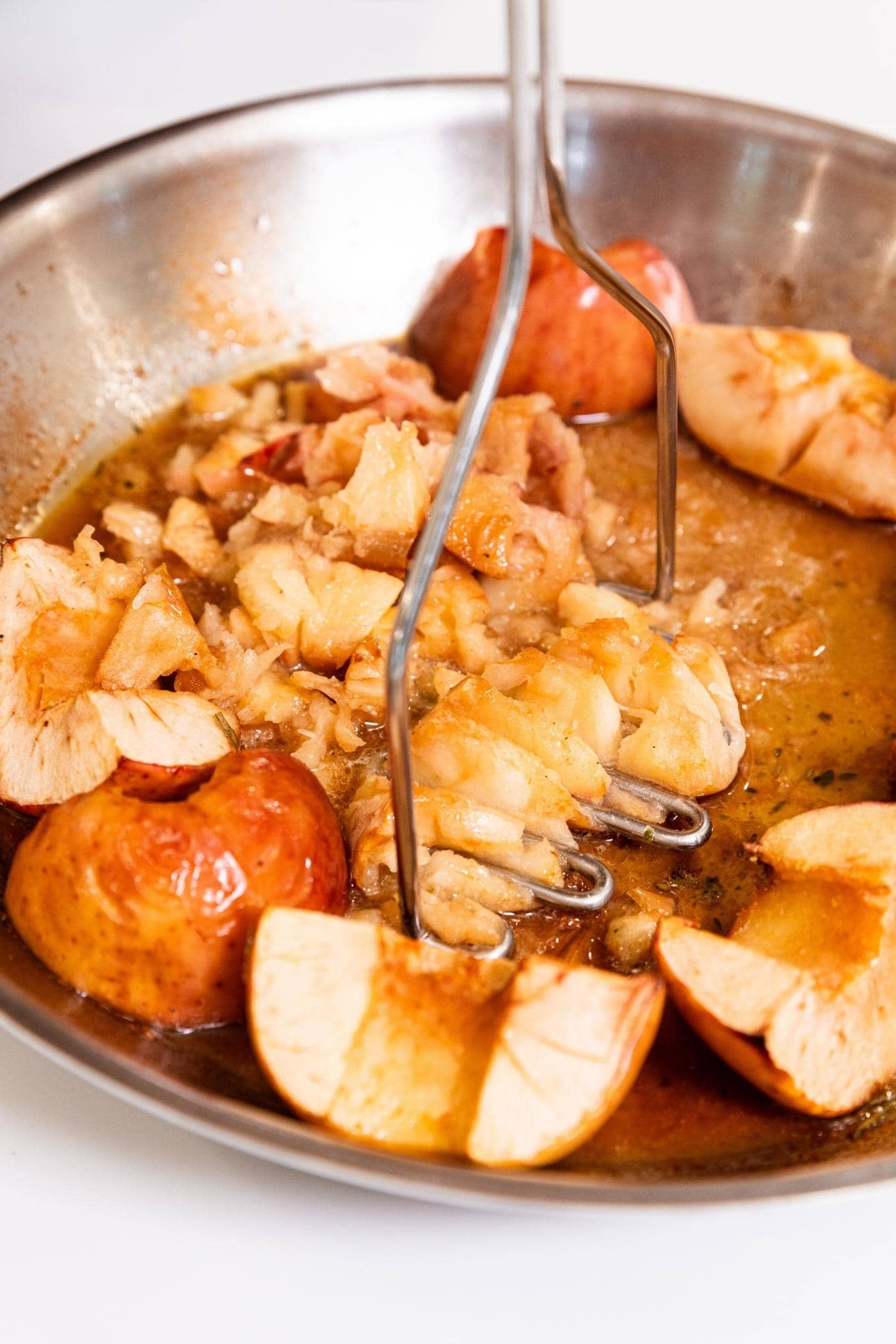 Masher smooshing apples into rough applesauce after being cooked in a skillet in the oven with pan juices collected at the bottom.