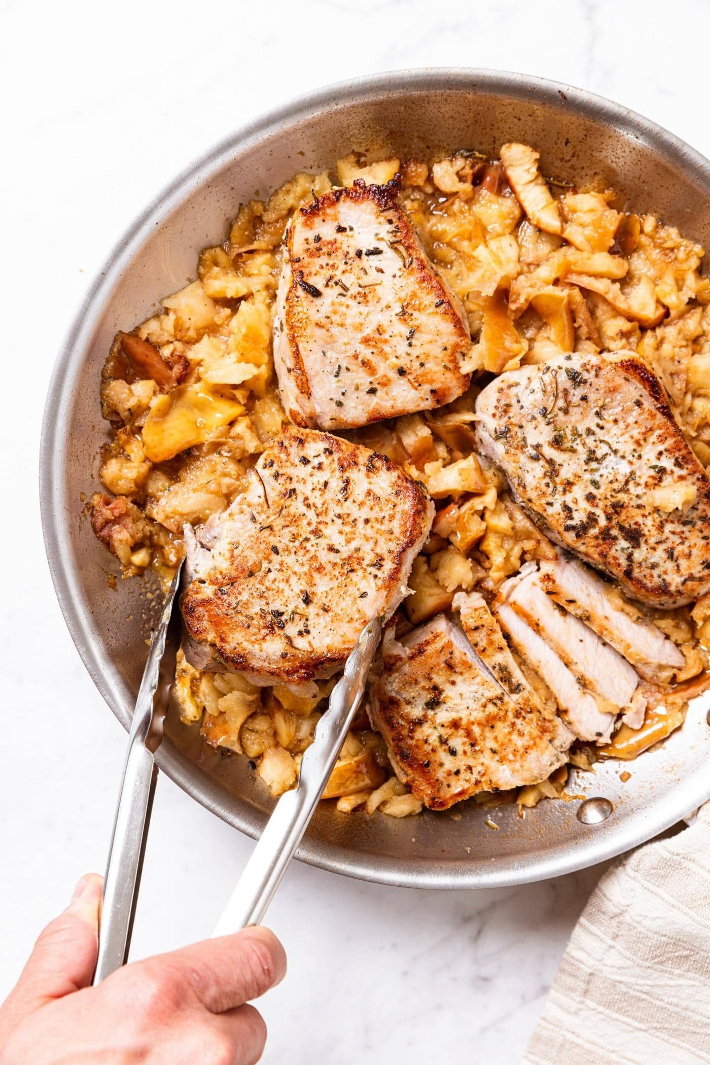 Metal skillet sitting on marble surface filled with smashed apples with browned pork chops being nestled on the top.
