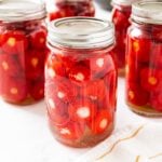 Glass jars filled with red stuffed pickled peppers sitting on white marble surface with towel to the side.