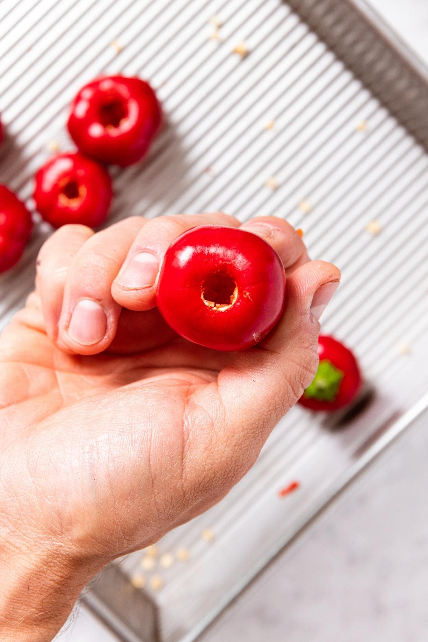 Hand holding small red sweet pepper with hollowed out core with more peppers below in metal baking dish.