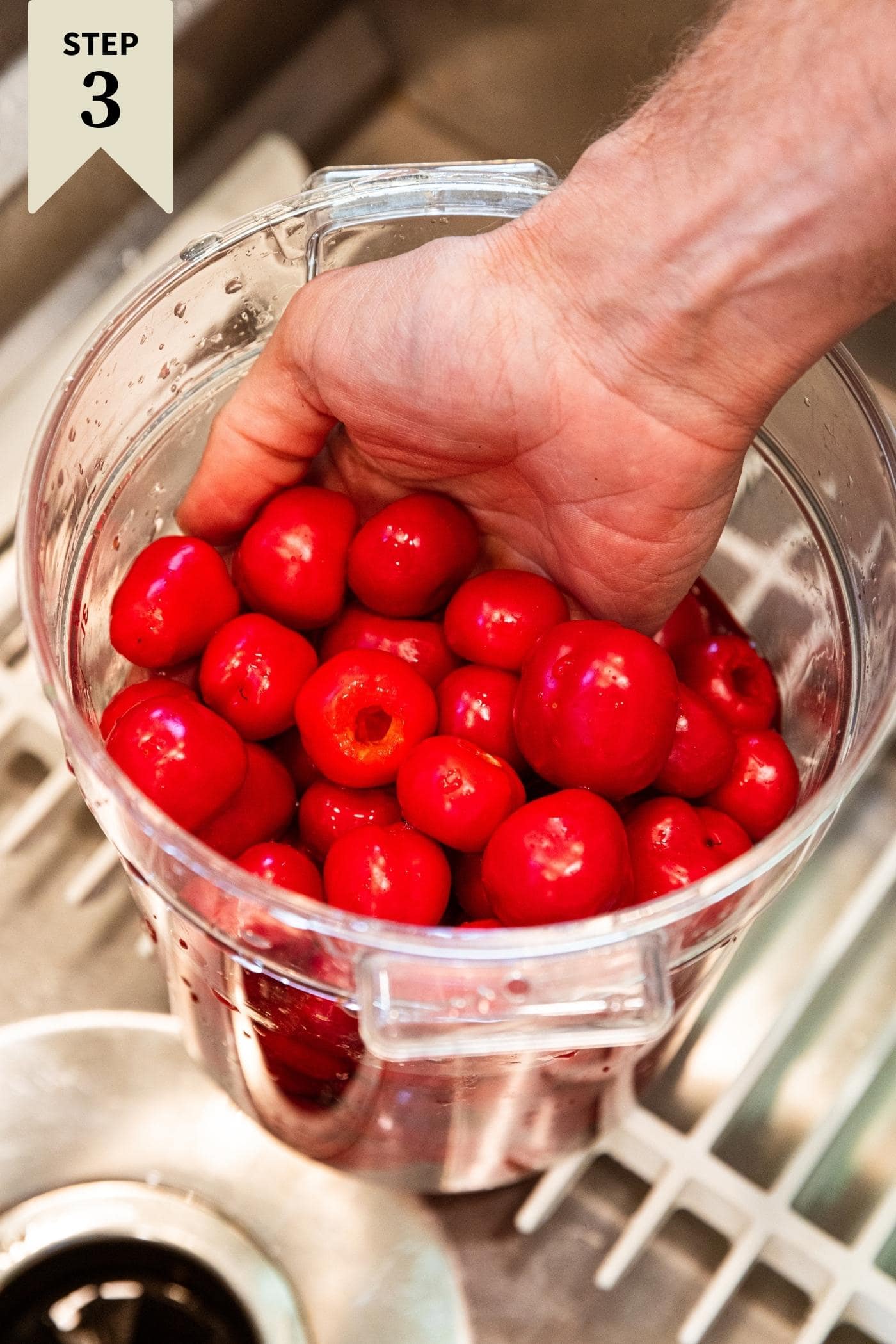 Hand scooping up small red sweet peppers sitting in plastic container filled with water in sink.