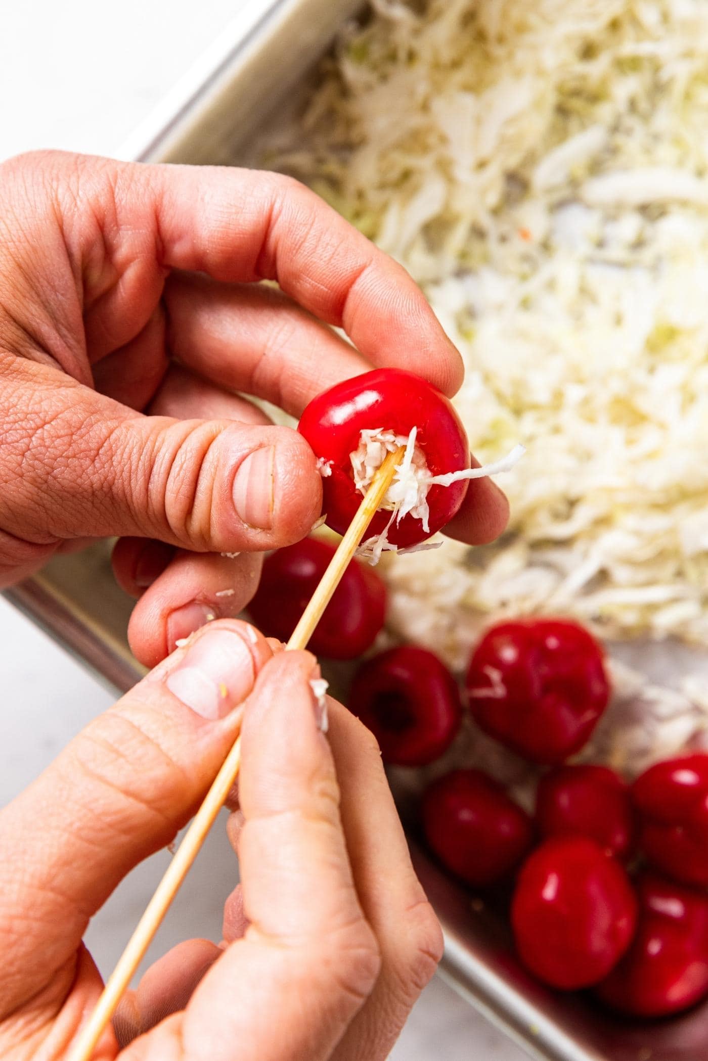 Two hands holding small red pepper stuffing raw shredded cabbage inside with a wooden skewer.
