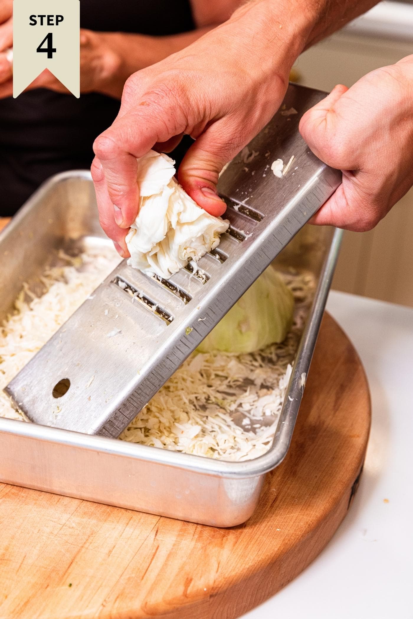 Hand holding shredder shredding raw cabbage across the surface with the cabbage collecting underneath in metal pan.