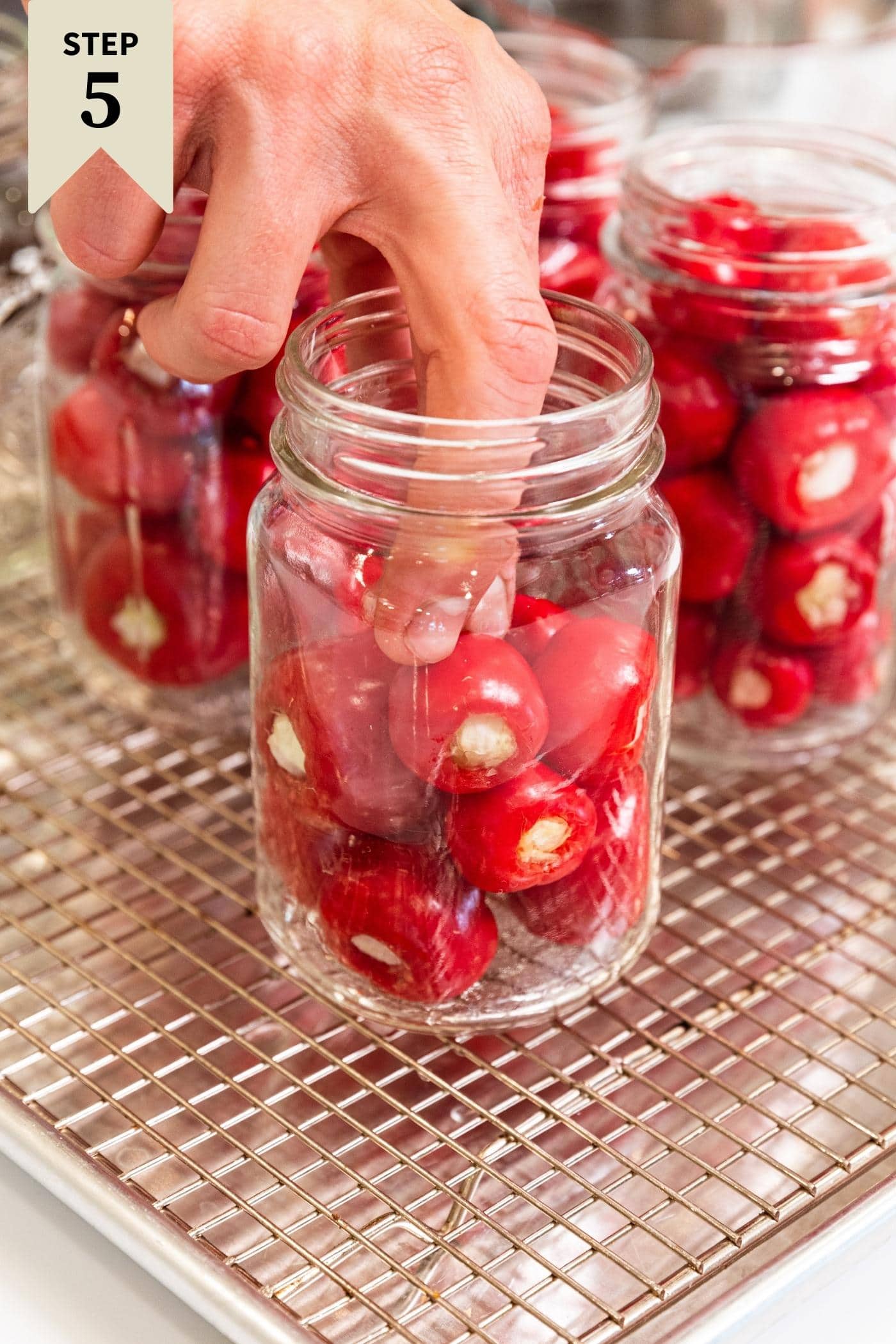 Hand placing small red stuffed peppers in glass canning jar on wire rack with more jars in background.