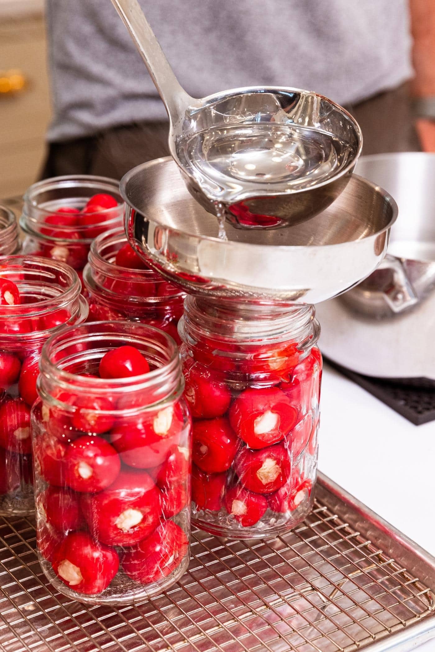 Metal ladle pouring clear brine liquid into glass canning jars with metal funnel on top of jar to keep the liquid going where desired.