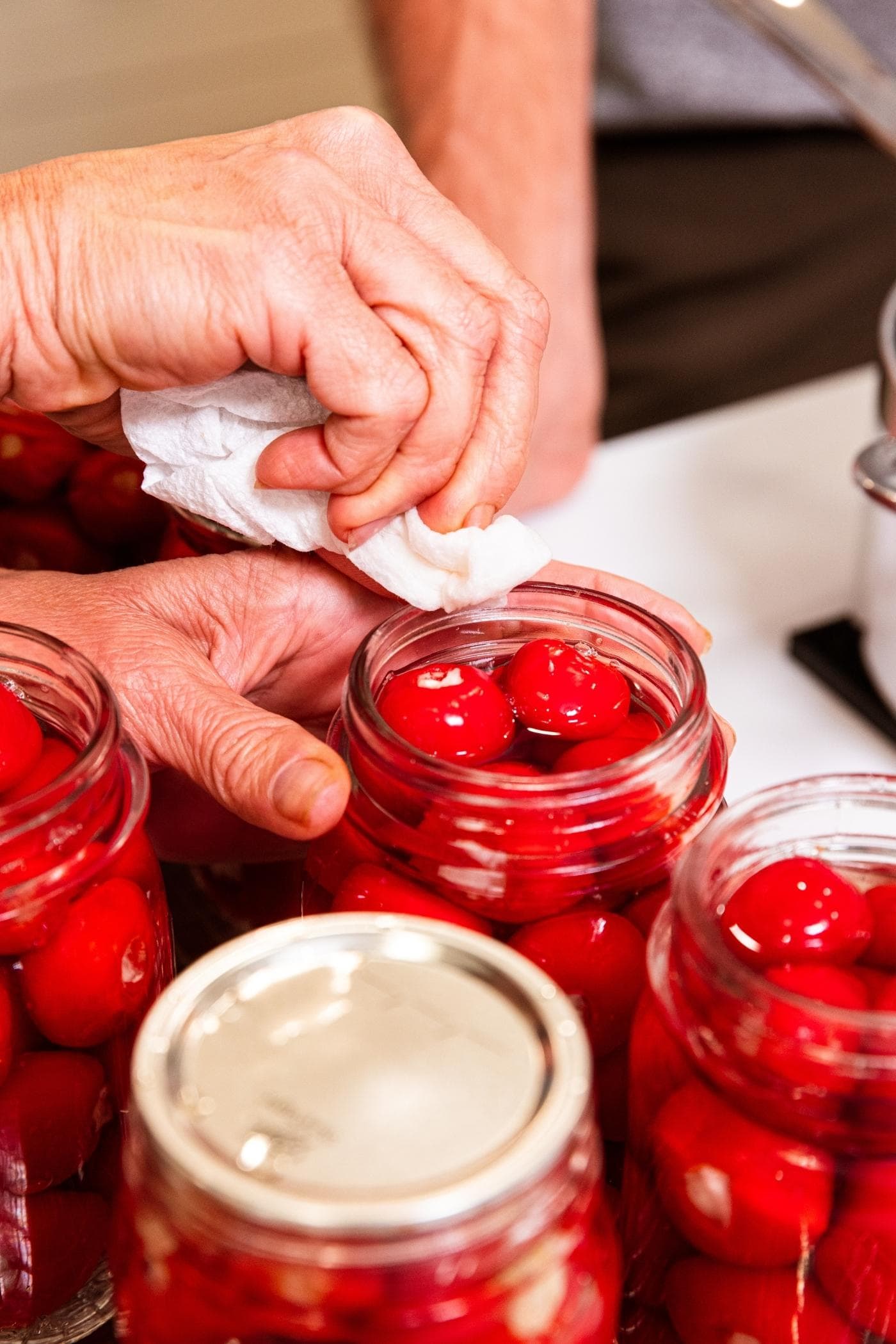 Hand using paper towel to wipe off the rim of glass canning jar filled with small red stuffed peppers.