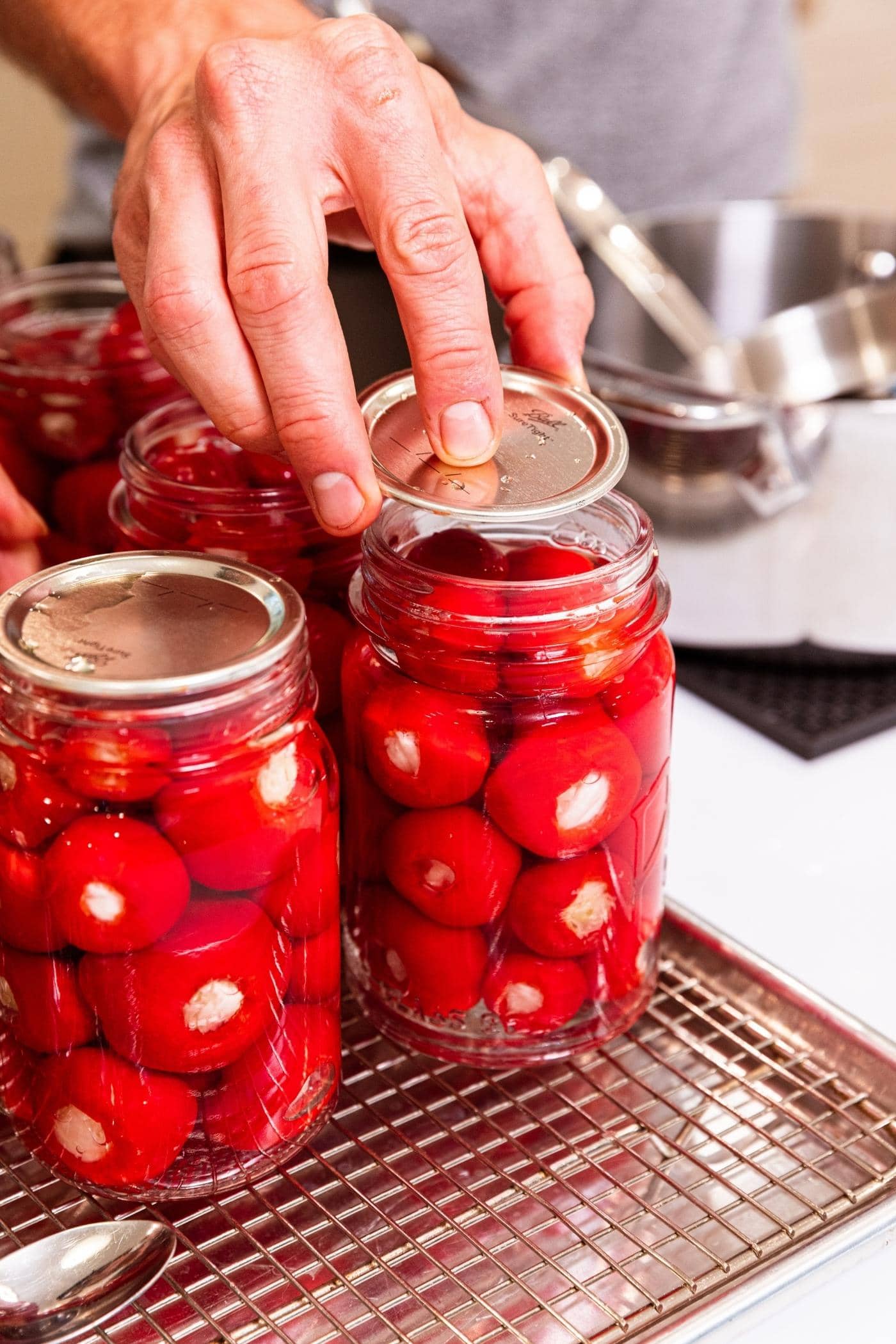 Hand placing lid on top of glass canning jar filled with stuffed red sweet peppers with jars sitting on metal rack.