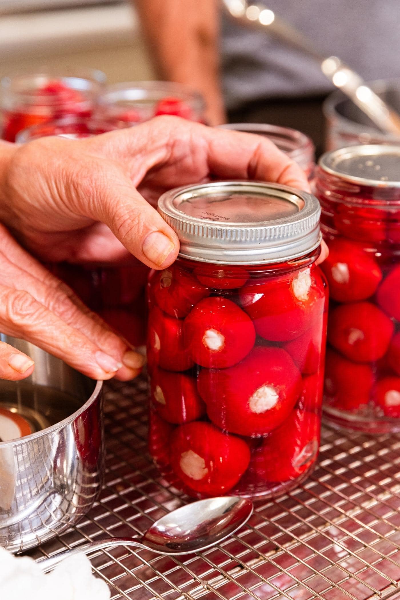 Two hands screwing on the ring to a canning jar packed with stuffed small red peppers with metal rack underneath.