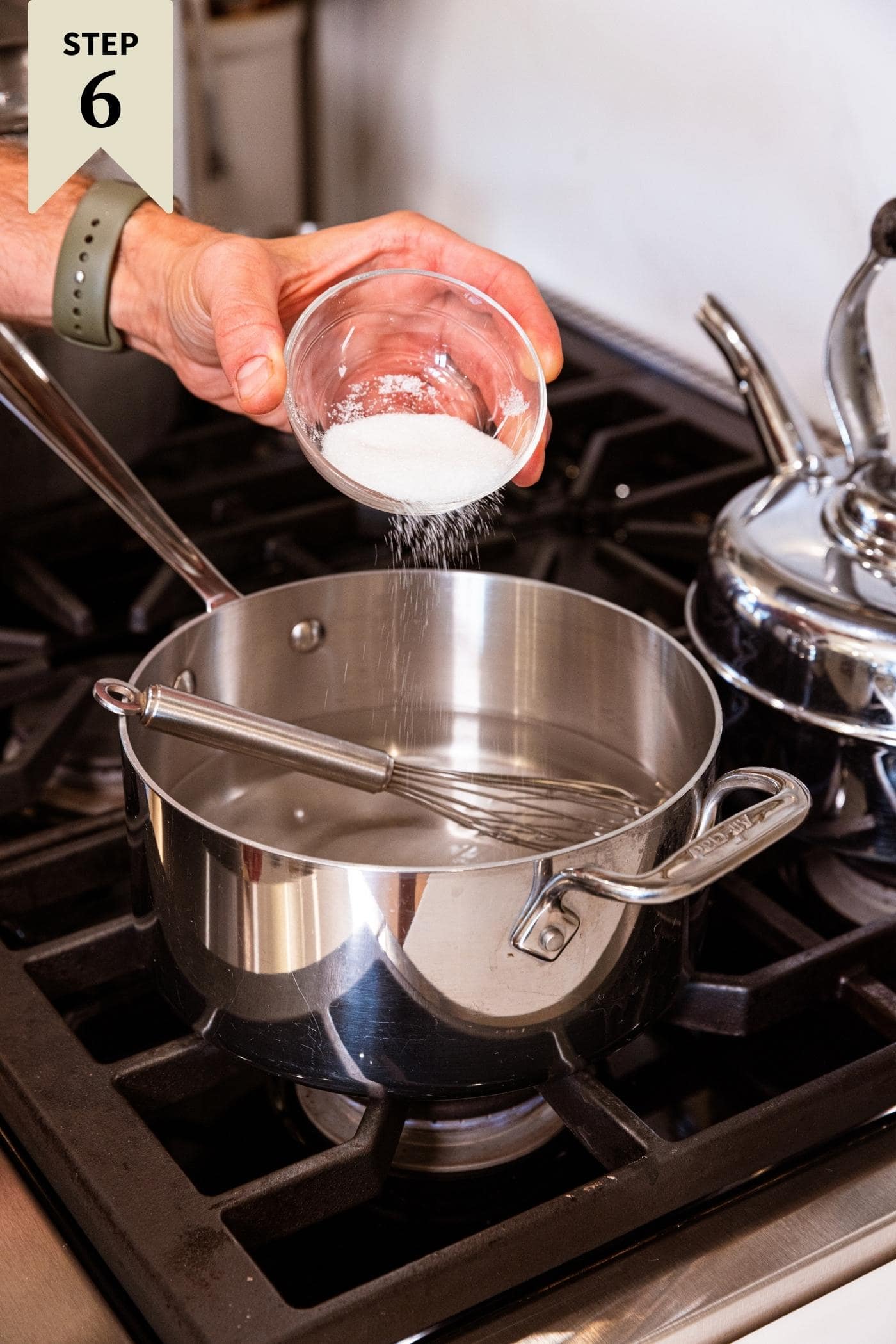 Hand pouring salt into clear liquid in metal kettle on stovetop.