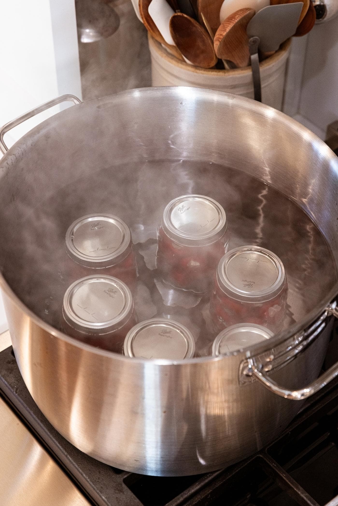 Large metal pot sitting on stovetop filled with water with six glass canning jars inside with steam coming off the top of the water bath.