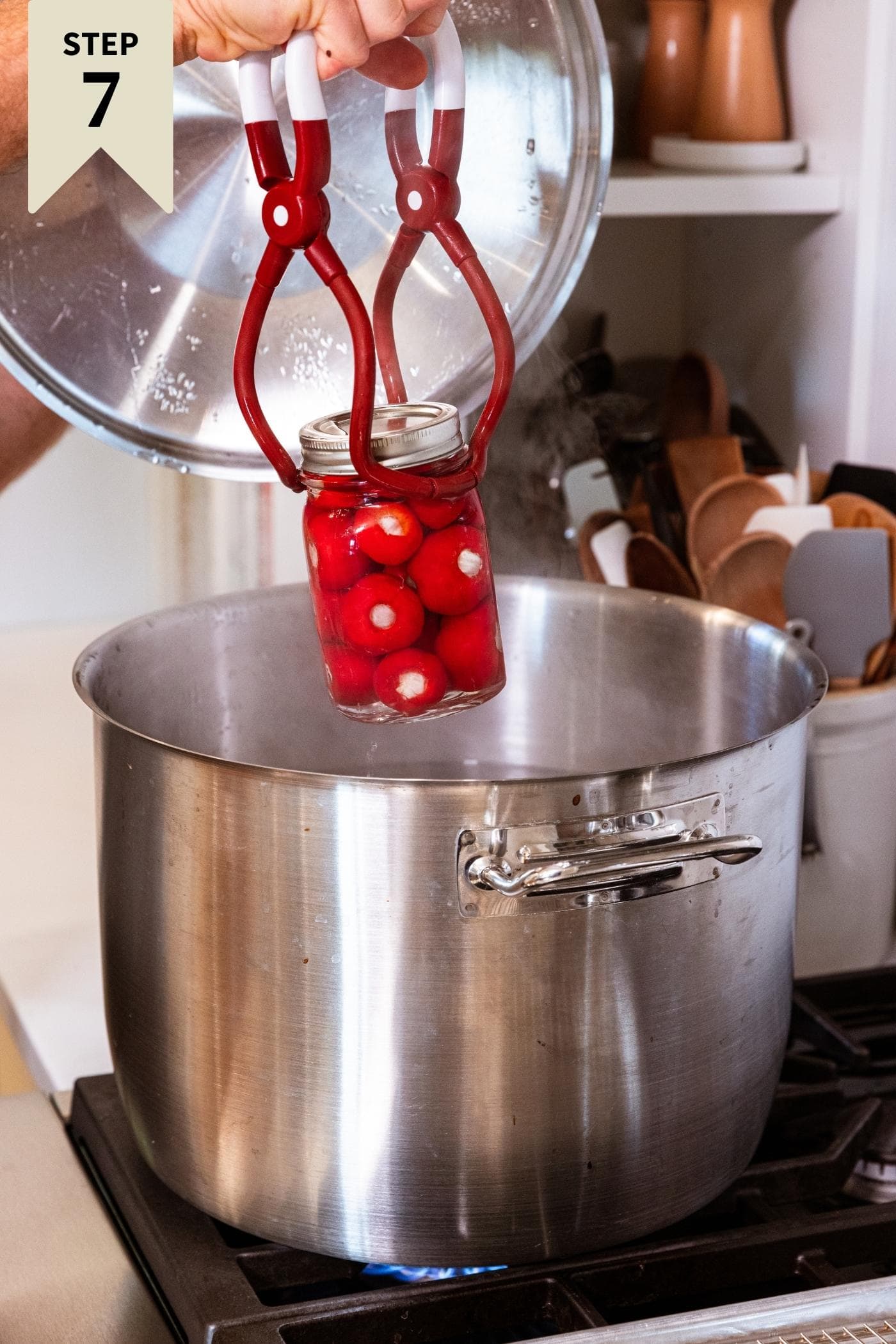 Hand using plastic tongs to place glass canning jar filled with stuffed red peppers into large metal pot to water bath the jars.