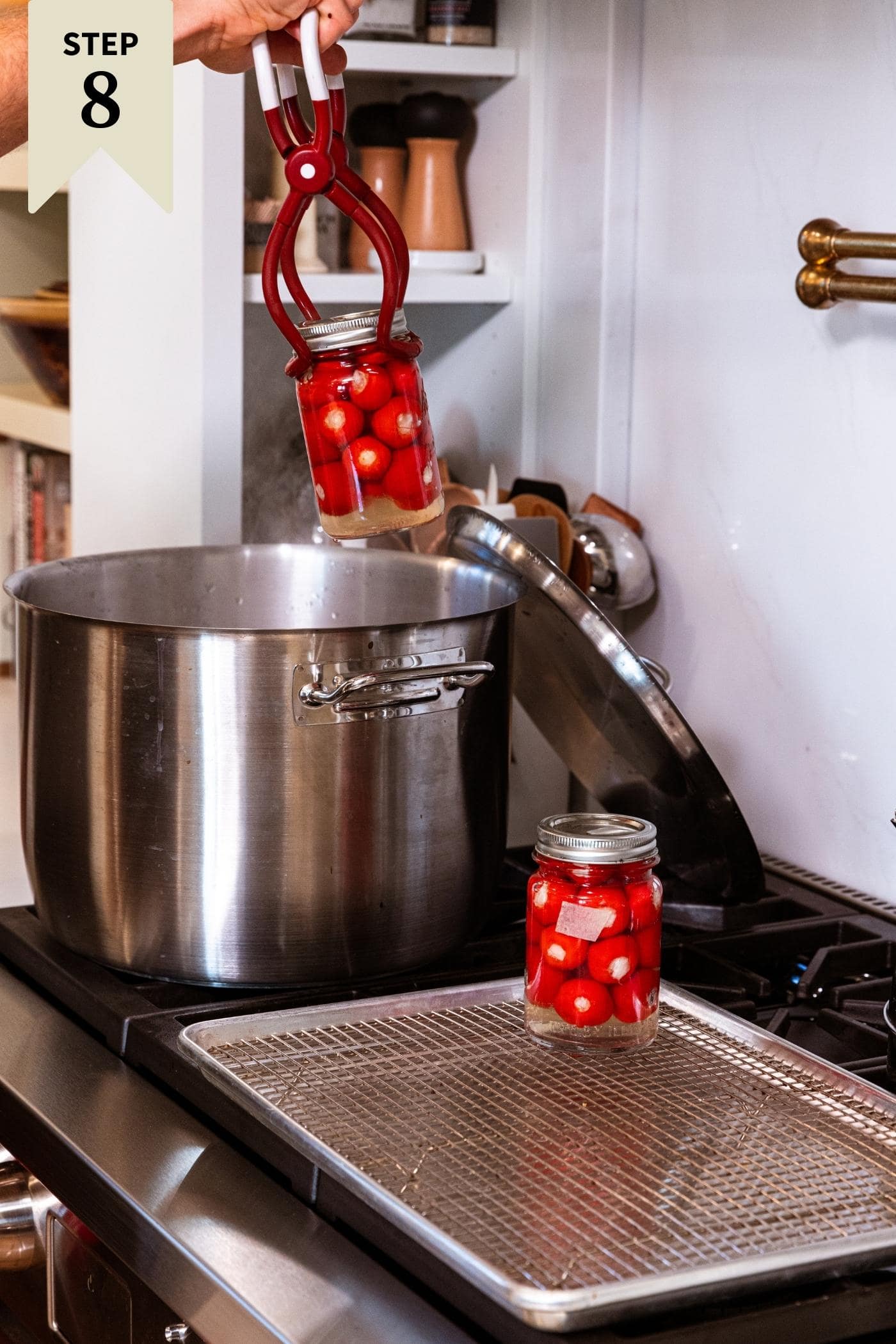 Hand pulling hot glass canning jars from water bath on stovetop in large metal pot with one jar already cooling on wire rack.