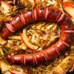 Large black cast iron skillet filled with cooked cabbage and apple with browned piece of sliced kielbasa on top all on marble surface.