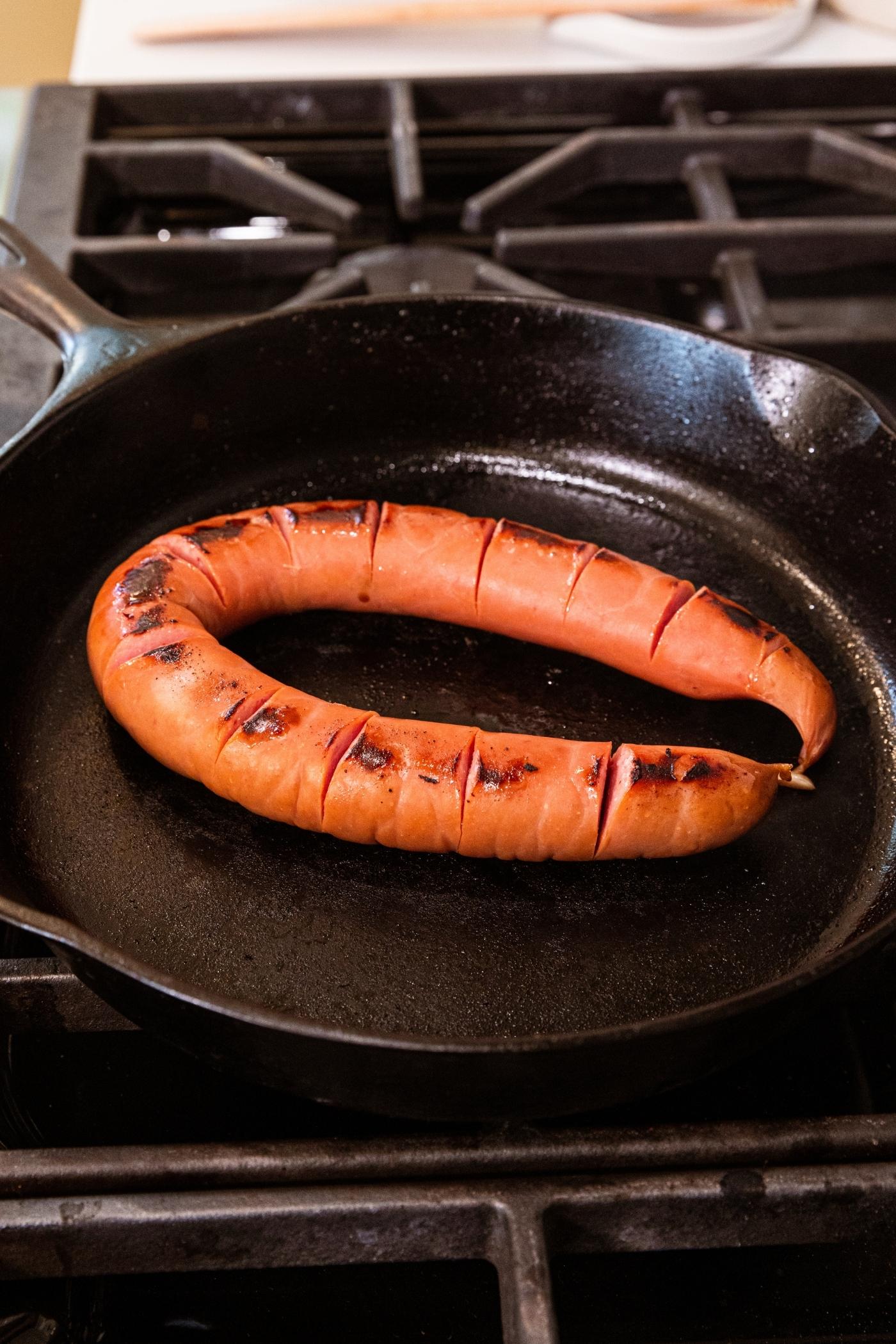 Black skillet with slightly browned piece of kielbasa sitting on heat all on stovetop.