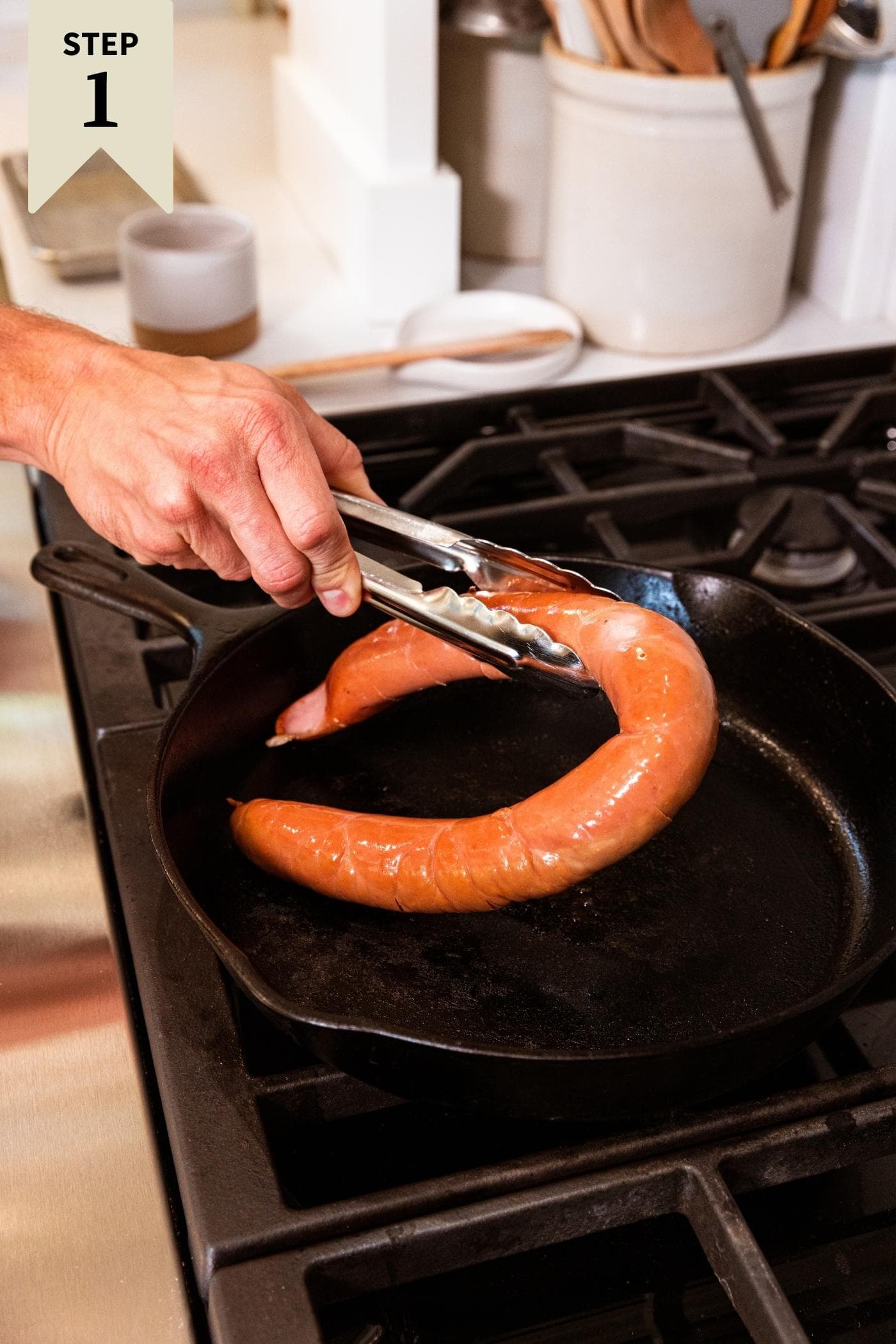 Hand holding tongs placing uncooked piece of kielbasa on black skillet on stovetop.