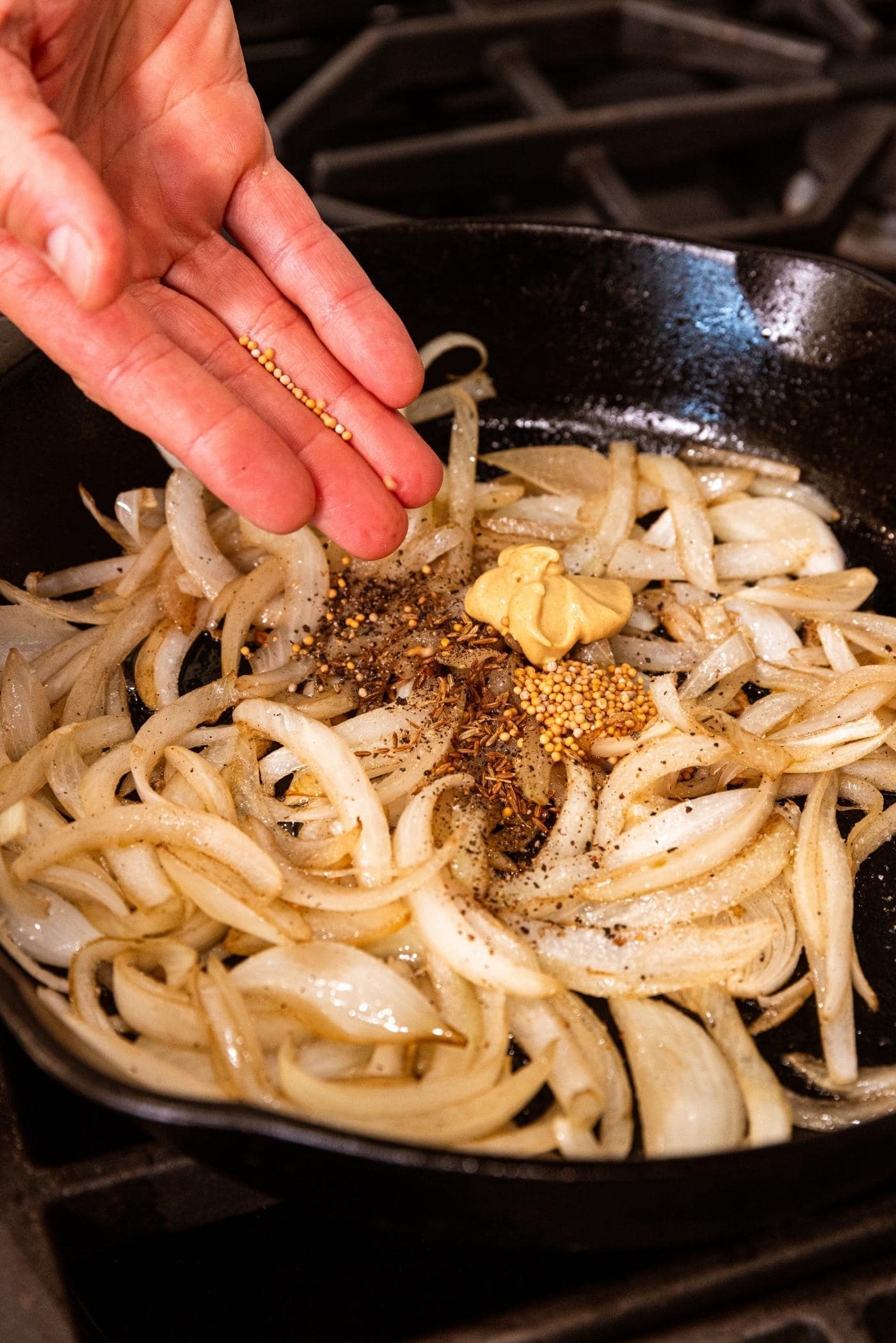 Hand pouring spices on top of caramelized onions in black pan.