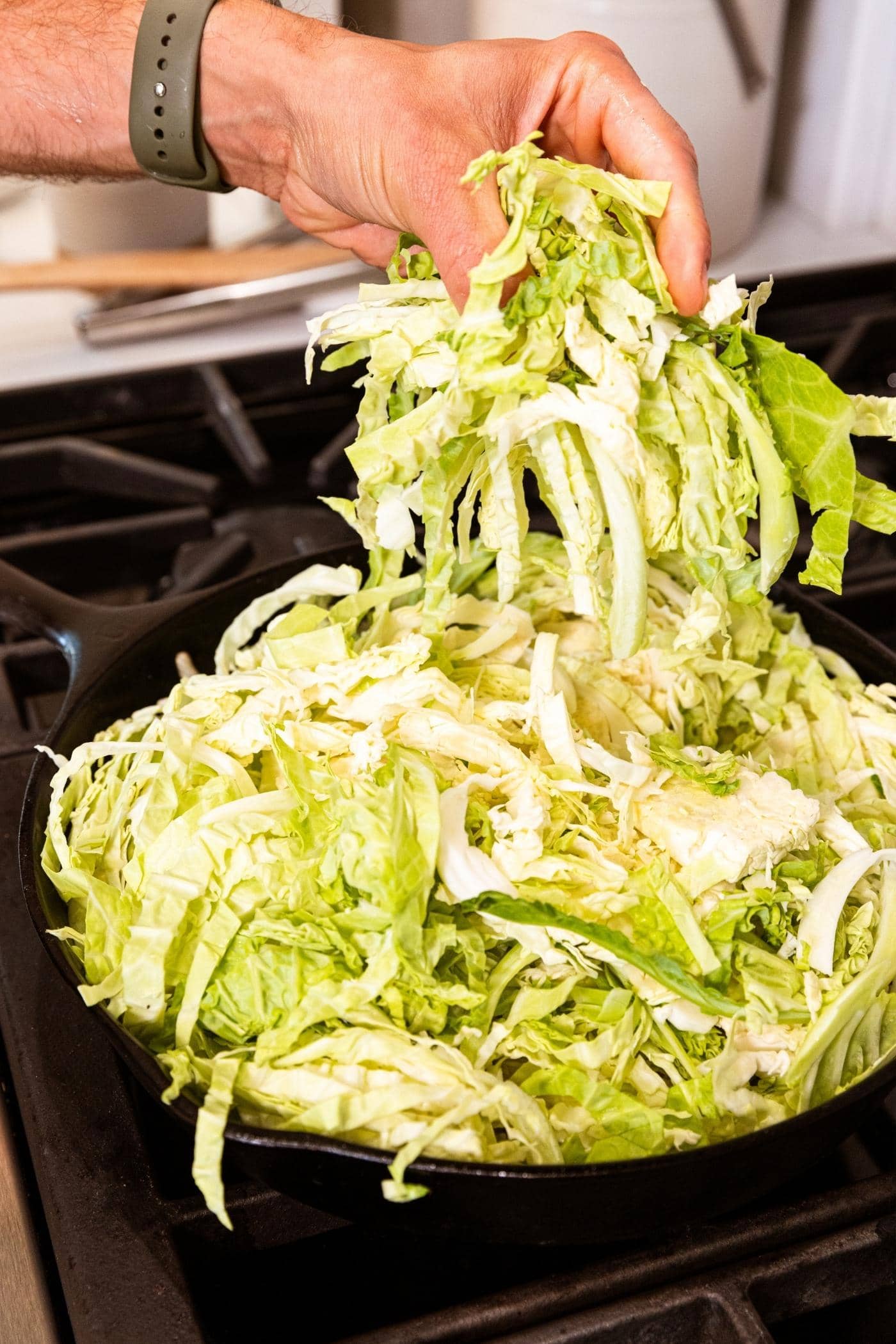 Hand dropping green and white colored cabbage into black cast iron skillet on stovetop.