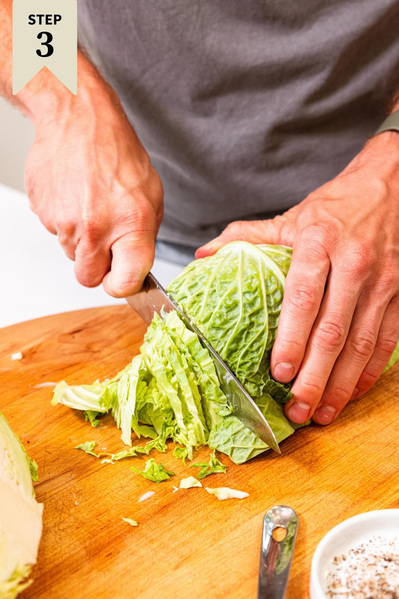 Hand holding green colored cabbage slicing into thin strips on wood cutting board.