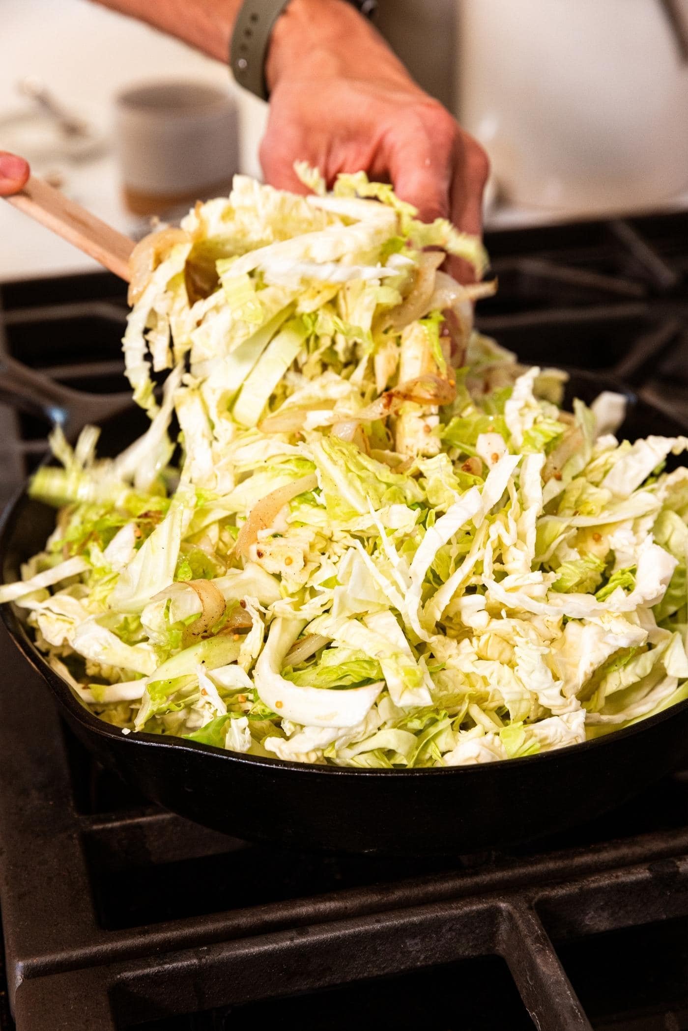Hand tossing shredded green and white colored cabbage in skillet on stovetop.