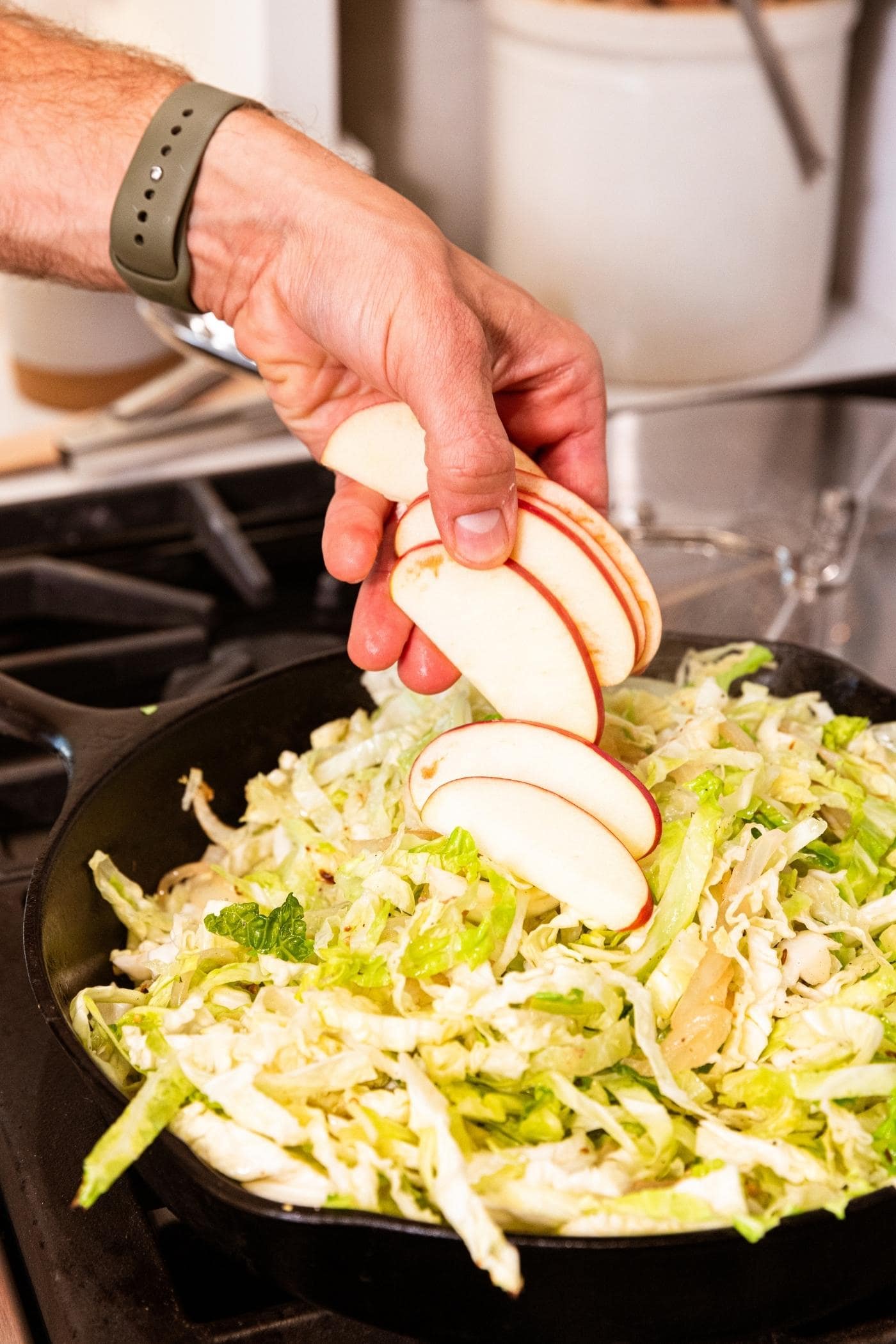 Hand dropping slices of apple on top of shredded cabbage in skillet on stovetop.