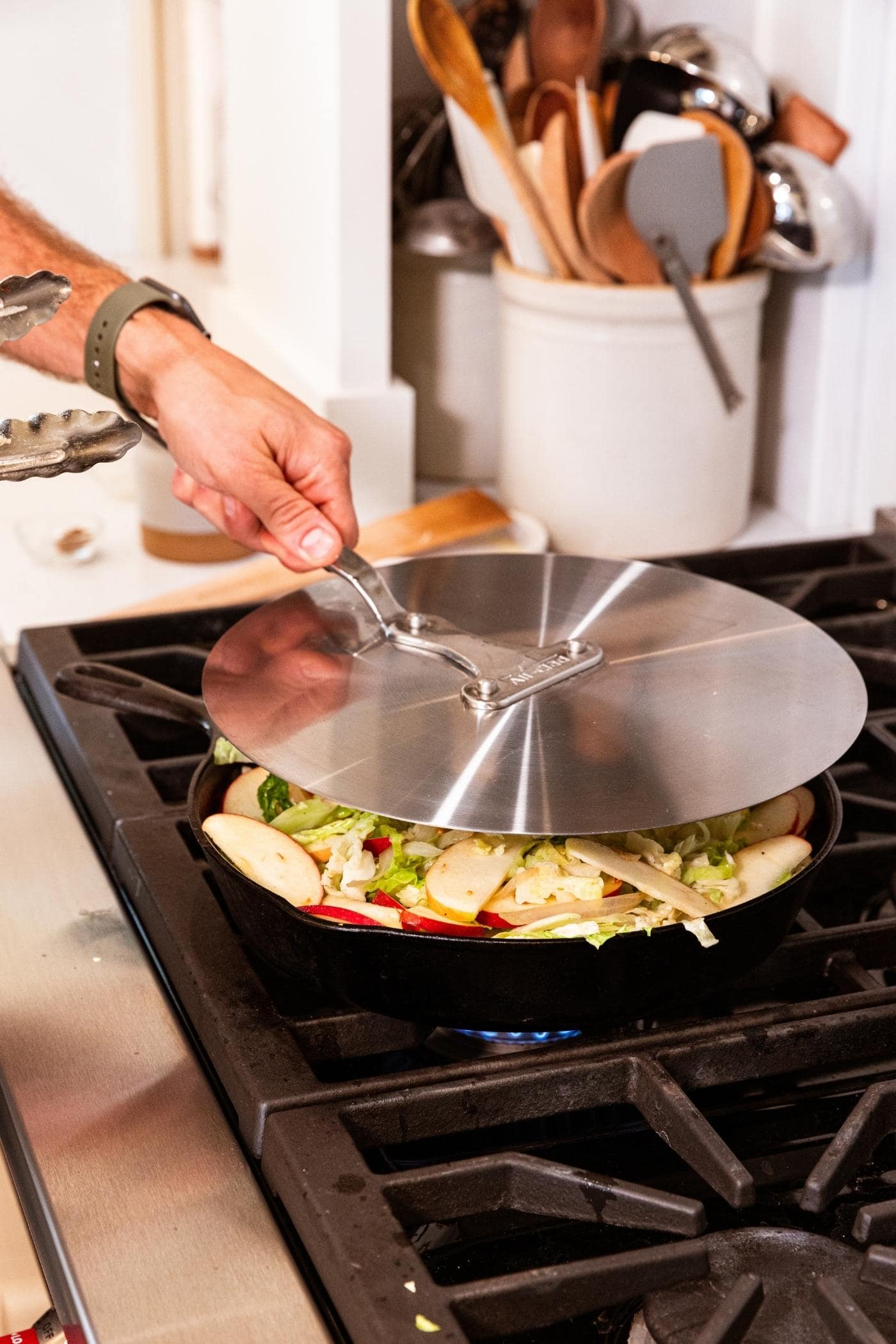 Hand placing metal cover on top of a black cast iron skillet on stovetop with shredded cabbage and apple inside.