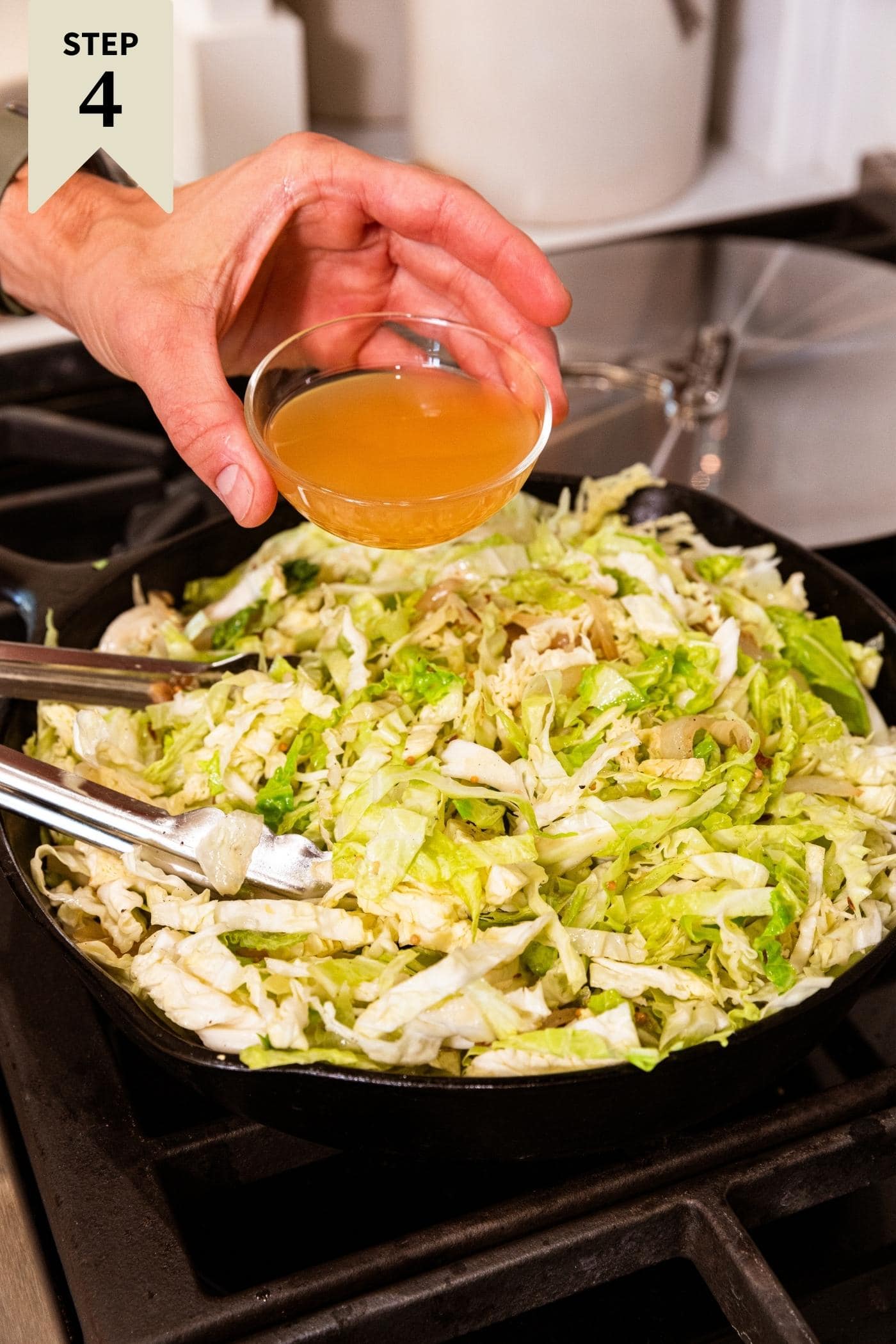 Hand pouring apple cider vinegar into cast iron skillet on stovetop from small glass bowl with shredded cabbage already in skillet.