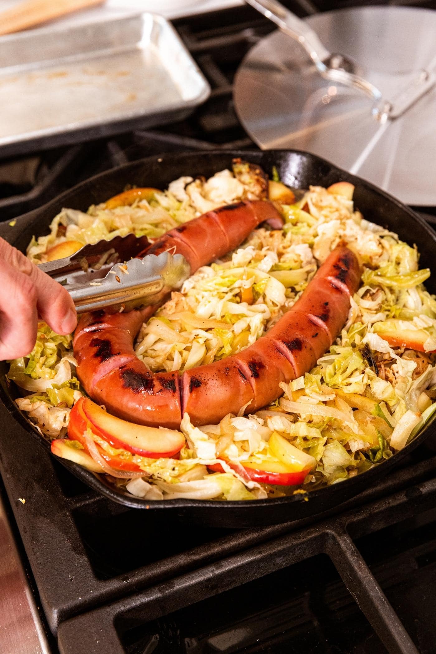 Tongs placing piece of kielbasa on top of cooked shredded cabbage and apple in skillet on stove.
