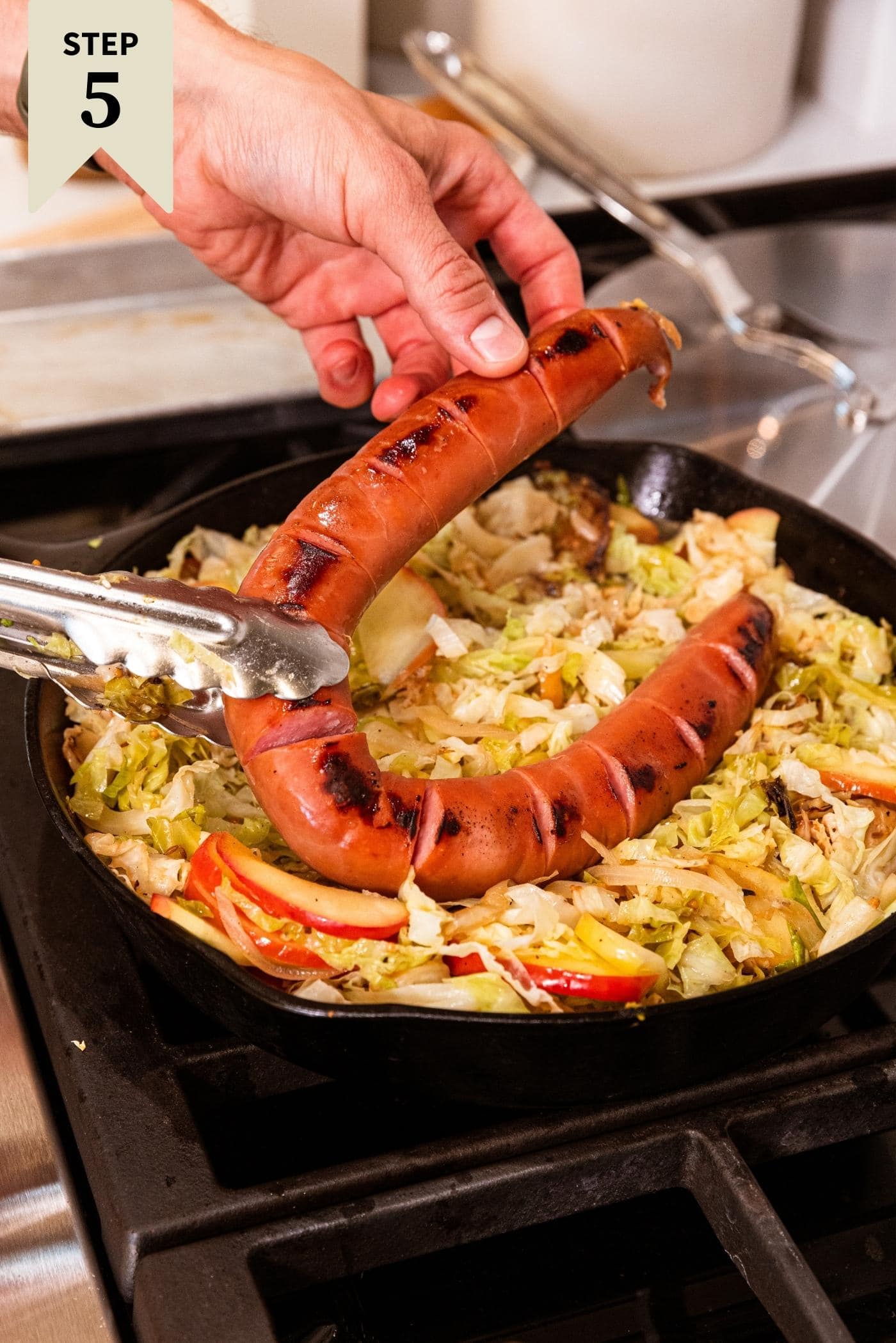 Hand placing large piece of browned kielbasa on top of cooked and shredded cabbage and slices of apple on stovetop in cast iron skillet.