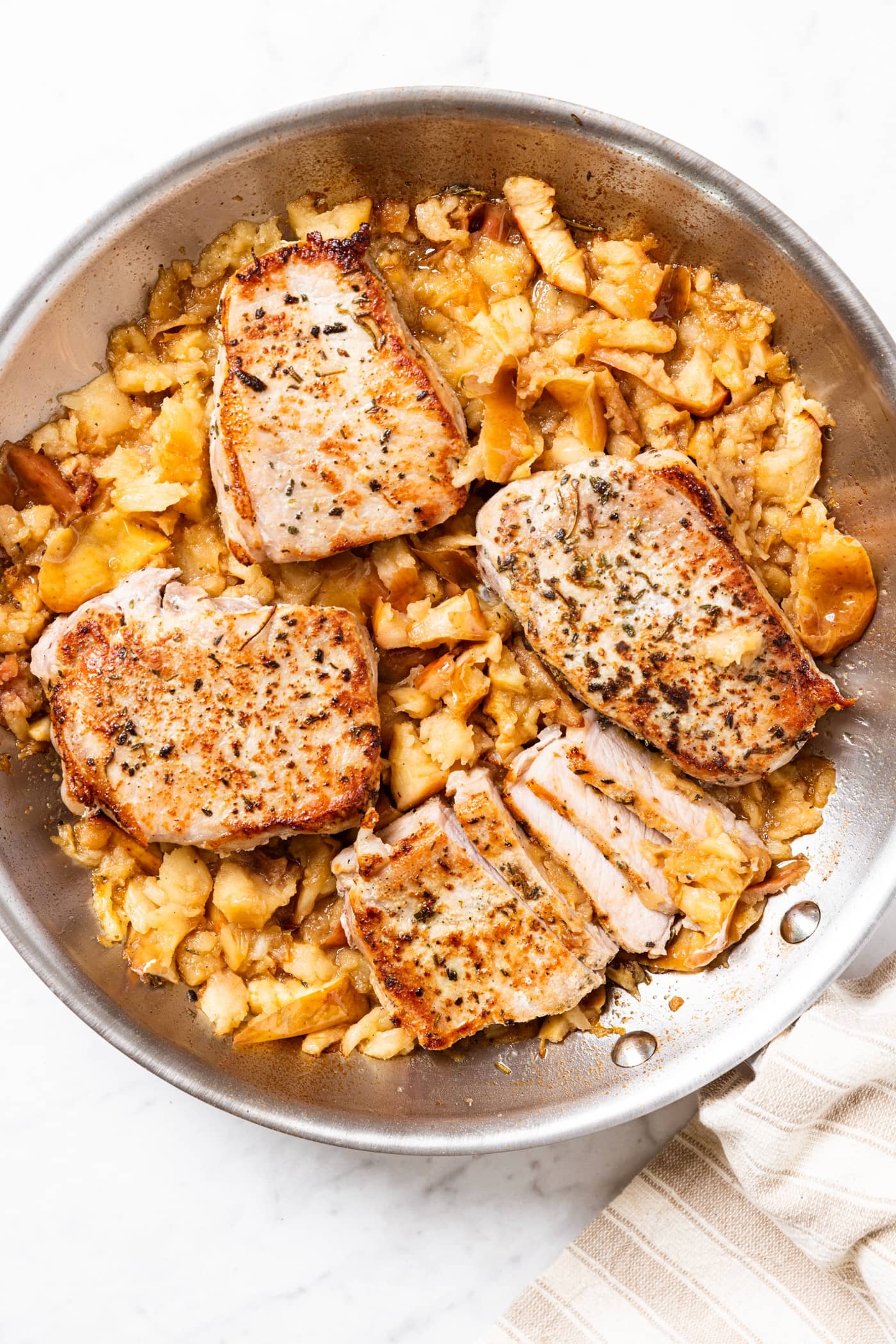 Metal skillet sitting on marble surface filled with smashed apples and four boneless pork chops nestled on top.