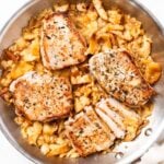 Large metal skillet sitting on white marble with cooked pork chops and mashed apples underneath right after coming out of the oven.