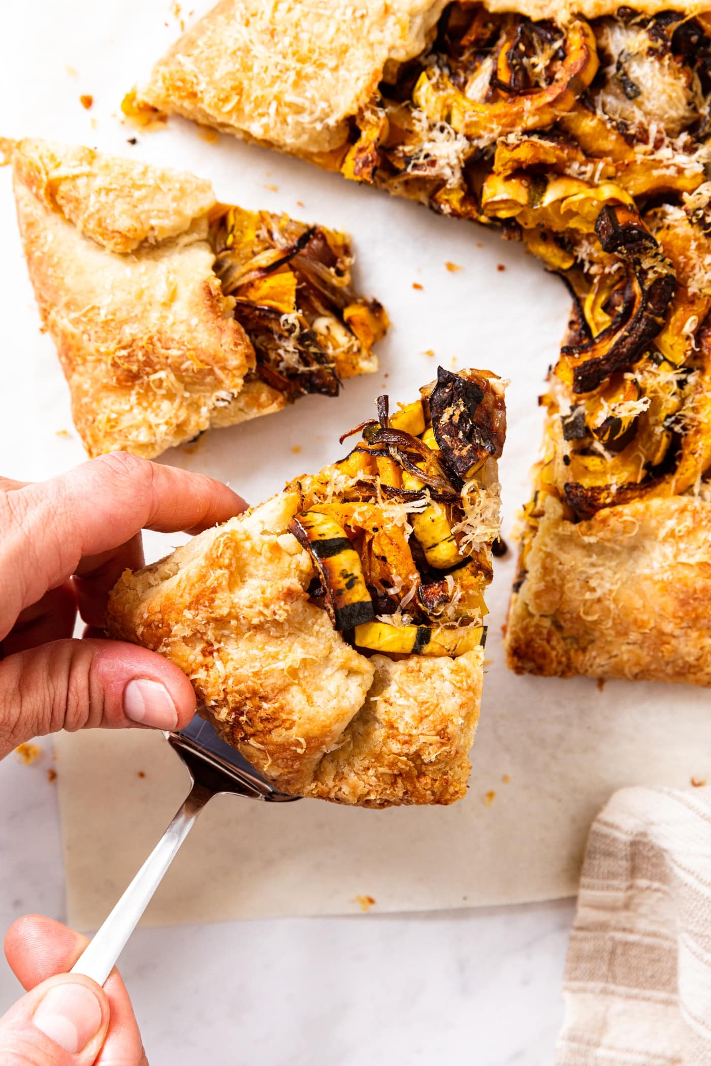 Hand holding pie server lifting up piece of savory roasted squash galette with rest of galette beside all on white piece of parchment.