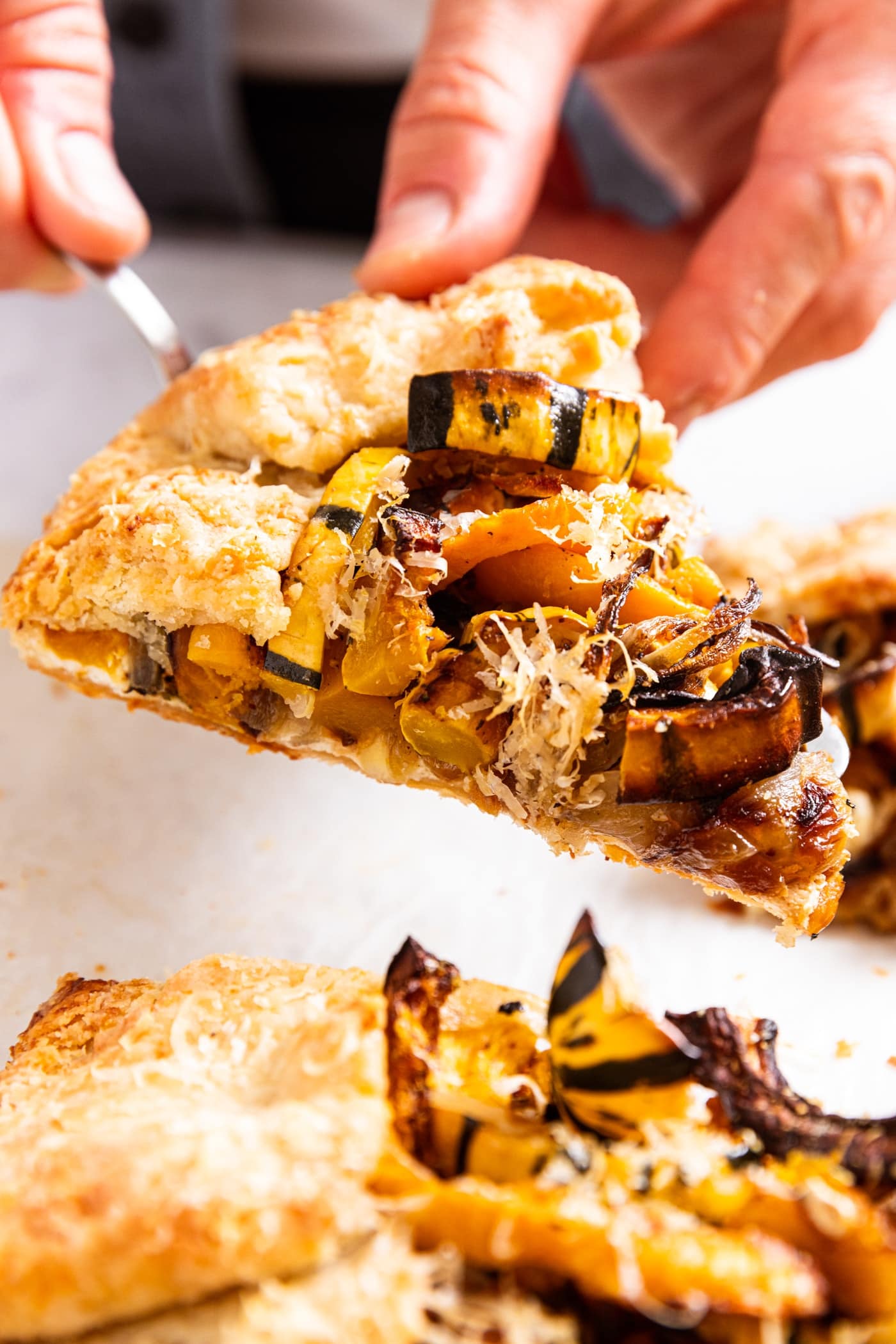 Two hands holding piece of roasted squash galette on pie serving utensil with rest of galette below.