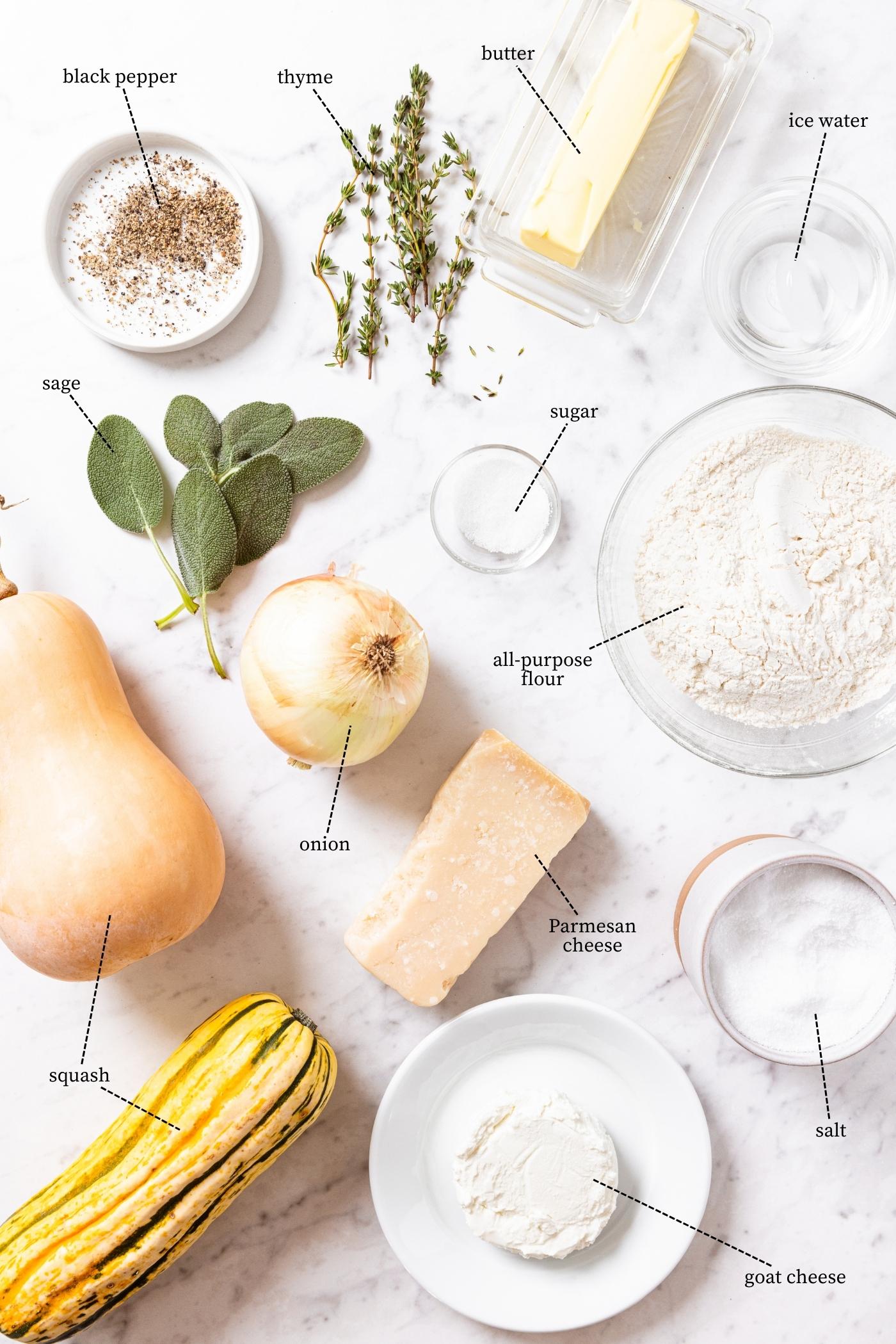 All ingredients needed to make savory roasted squash galette sitting on marble surface including squash, cheese, onion, herbs, salt, pepper, flour, and more.