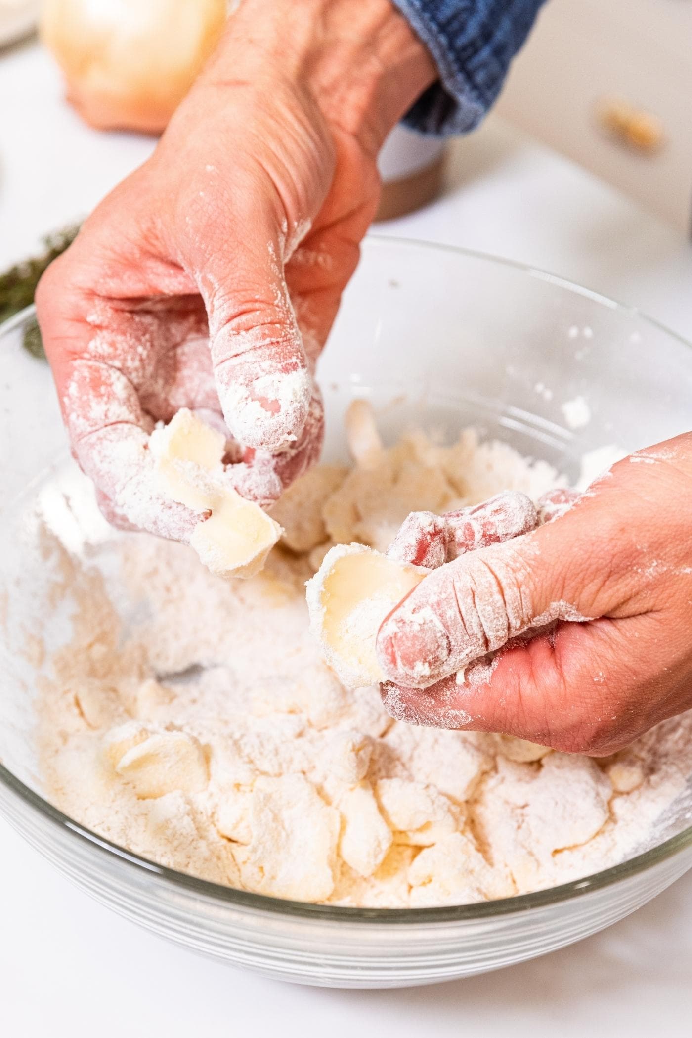 Two hands working butter into flour mixture in glass bowl sitting on white surface.