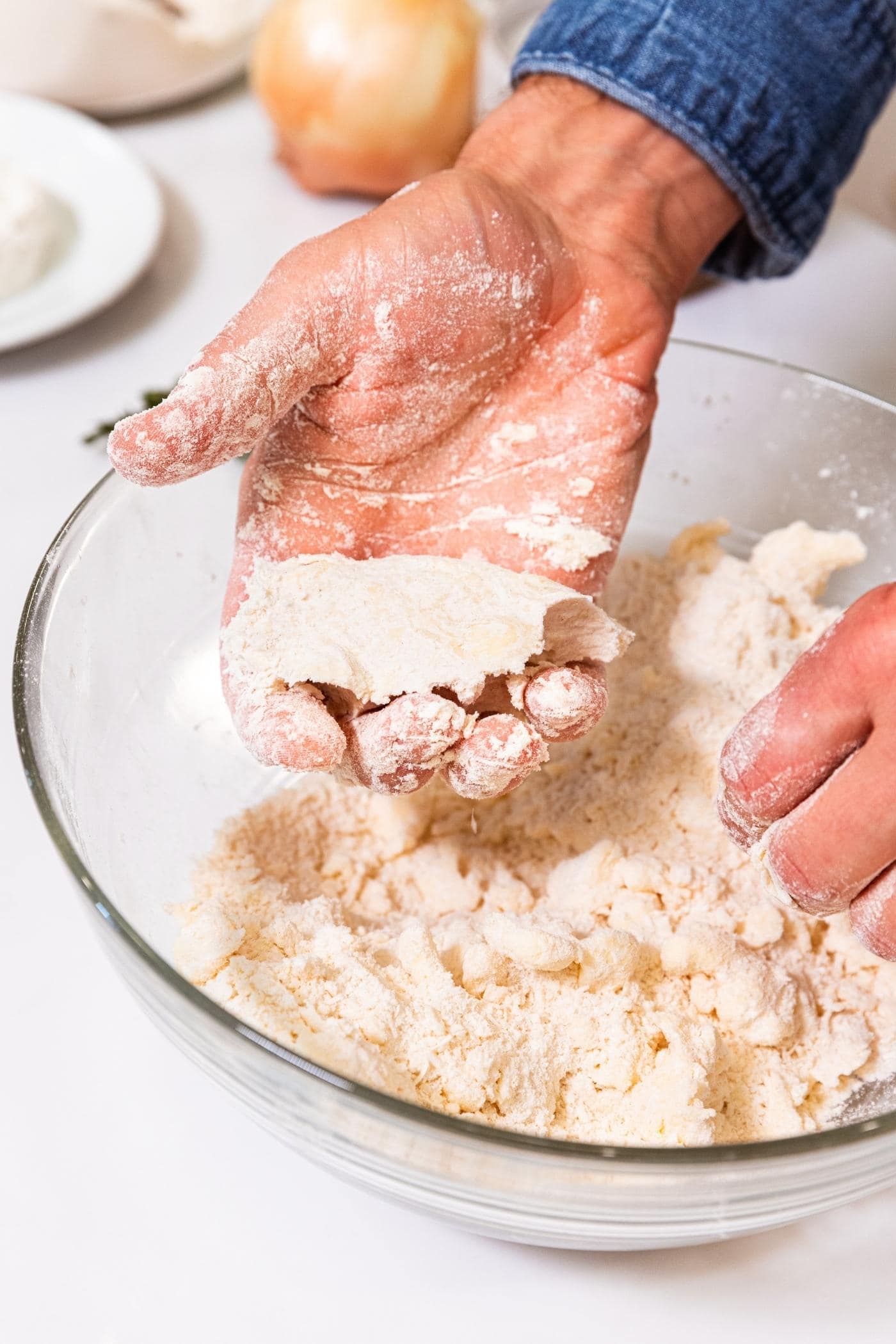 Hand holding fistful of dough after being pushed together with rest of dough in glass bowl below.