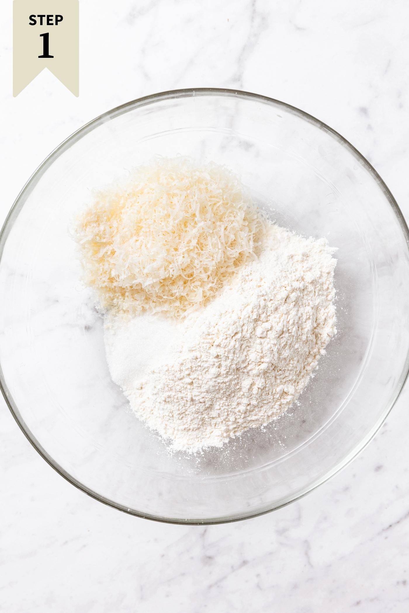 Glass bowl filled with flour, sugar, salt, and Parmesan cheese sitting on marble surface.
