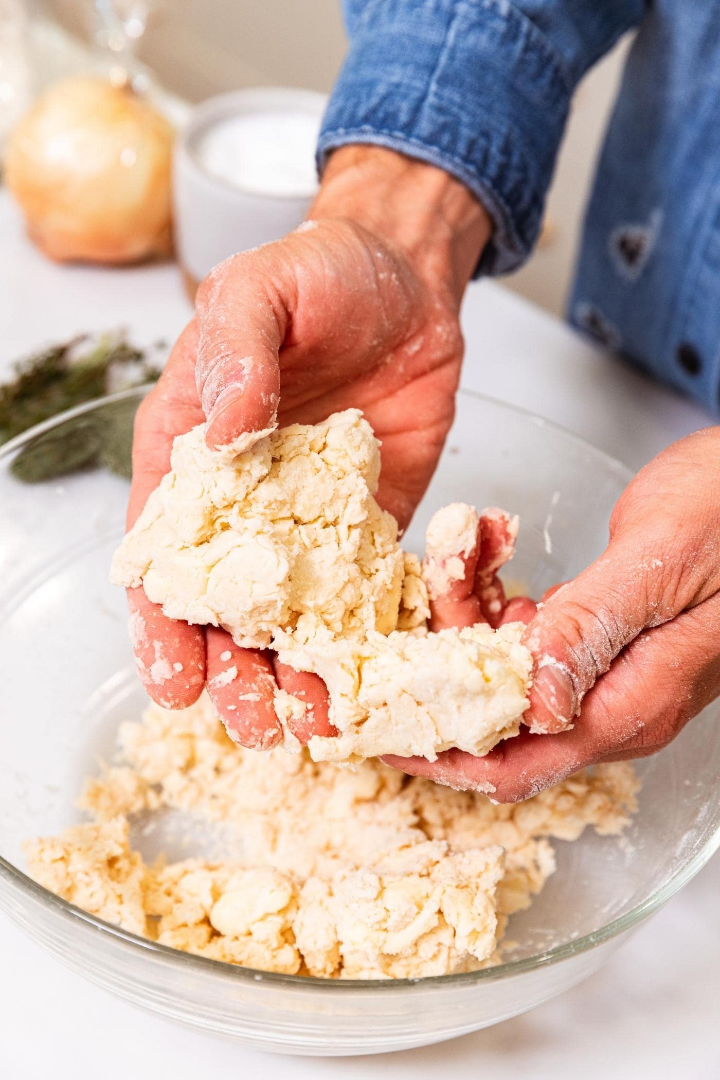 Two hands pulling apart compacted dough from glass bowl filled with remainder of dough on white surface with herbs and onion in background.