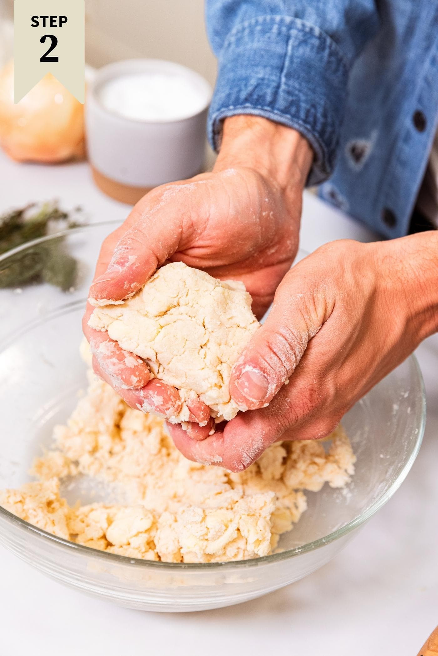 Two hands holding compacted piece of dough with rest of dough below in a glass bowl on white surface with green herbs in background.