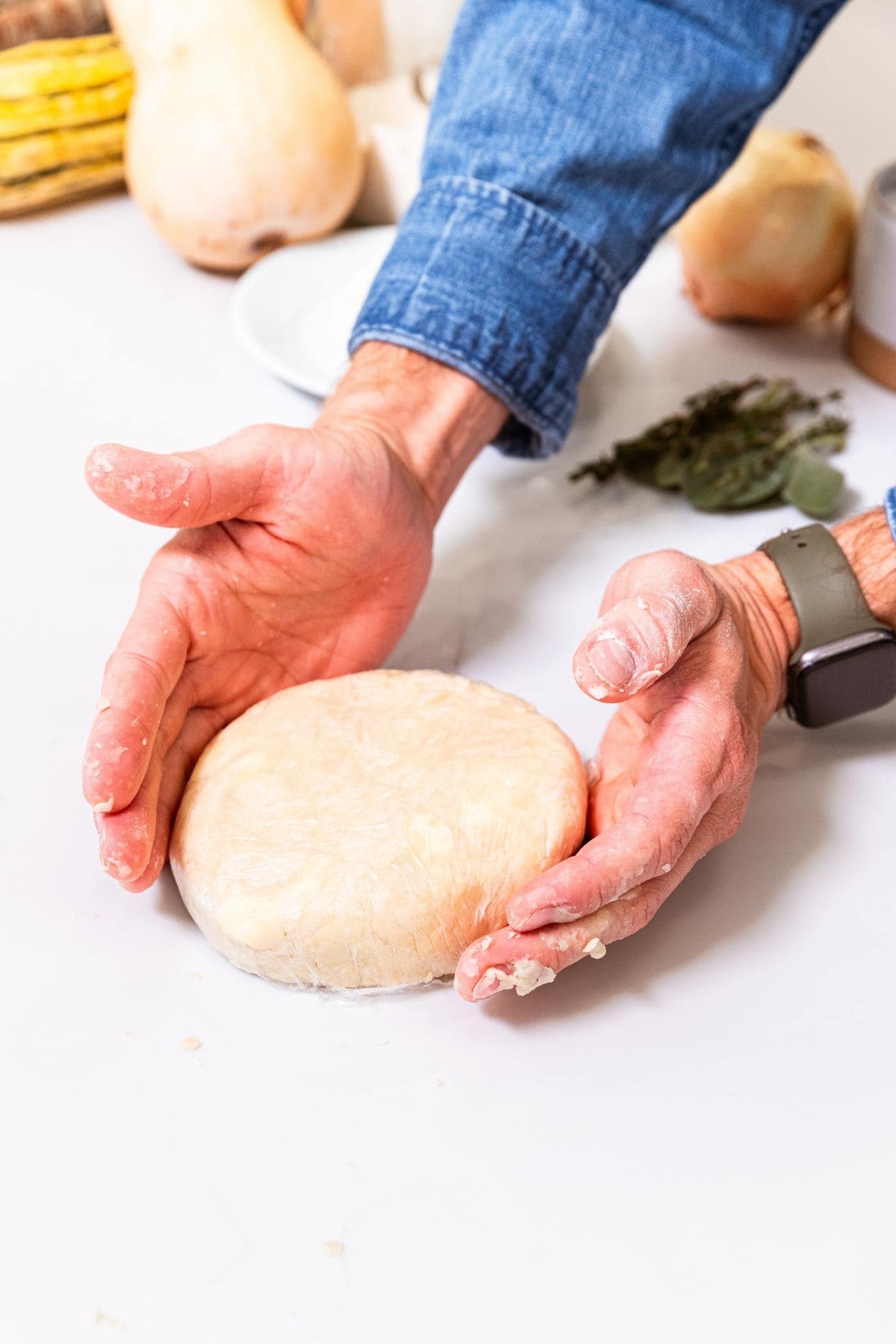 Two hands moving dough wrapped in plastic wrap around in circle on white surface with vegetables in background before making into a galette.