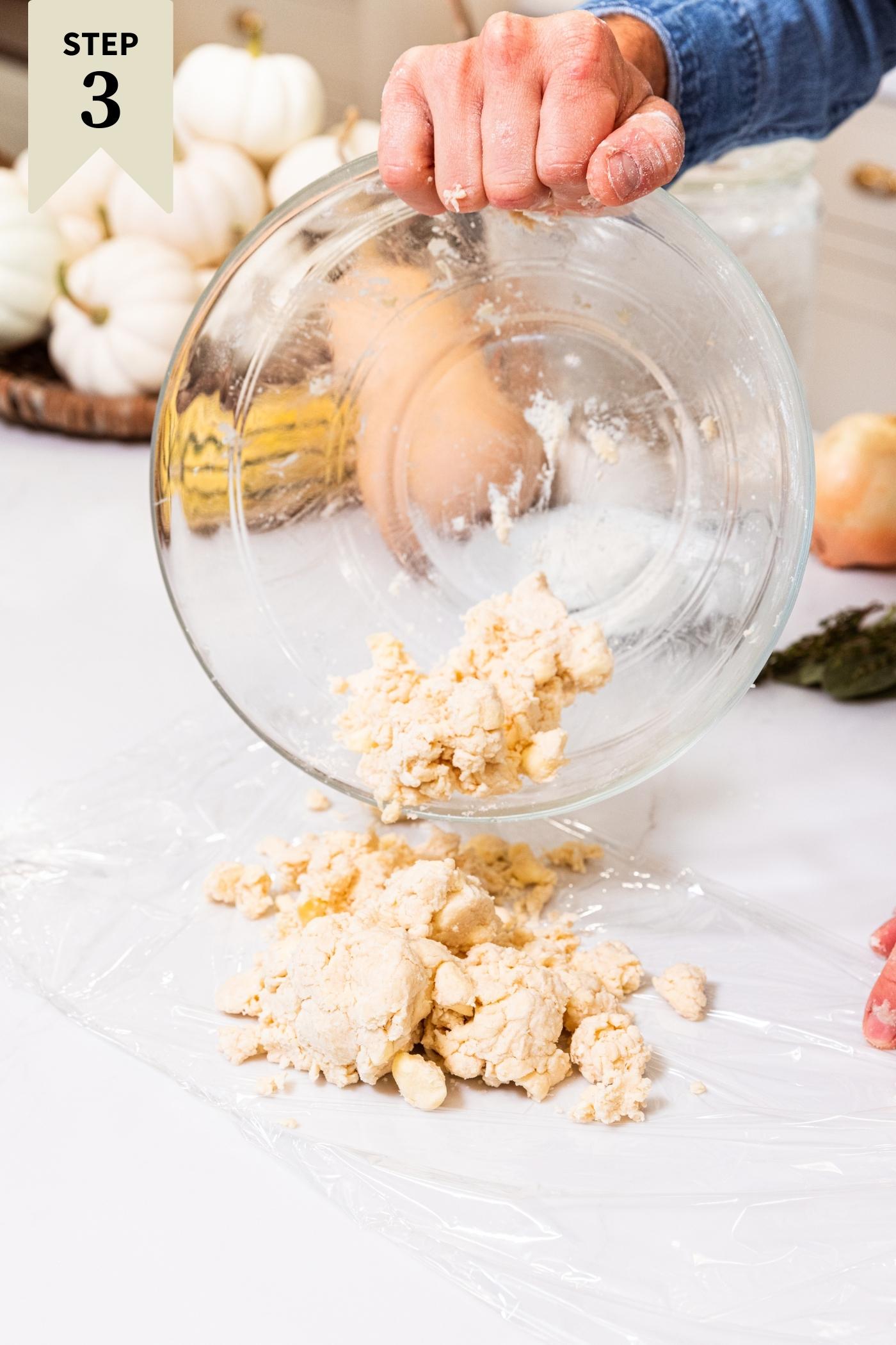 Hand dumping somewhat wet dough out of a glass bowl onto piece of plastic wrap on white surface.