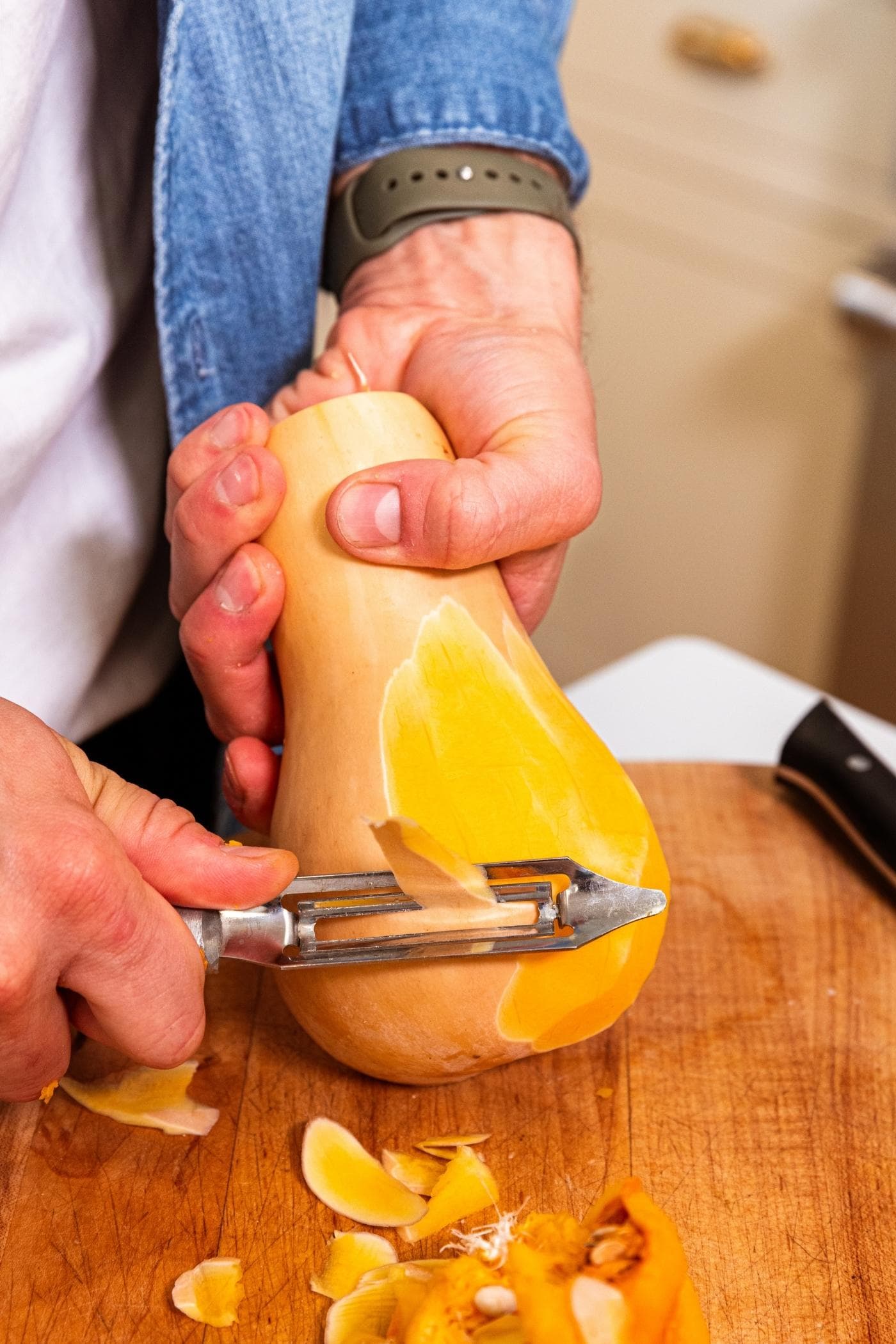 Hand holding butternut squash peeling the skin off with a peeler on wood cutting board.