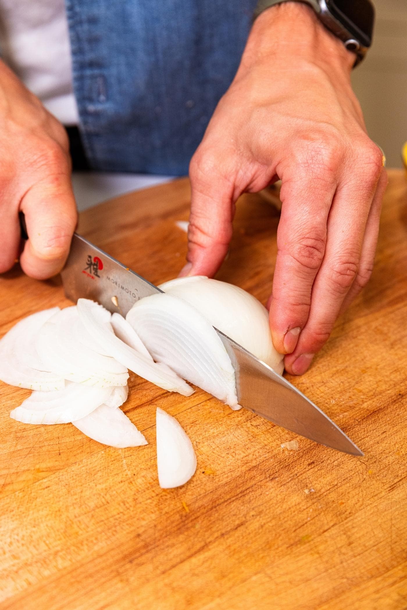 Hand slicing onion into small slices on wood cutting board in kitchen.