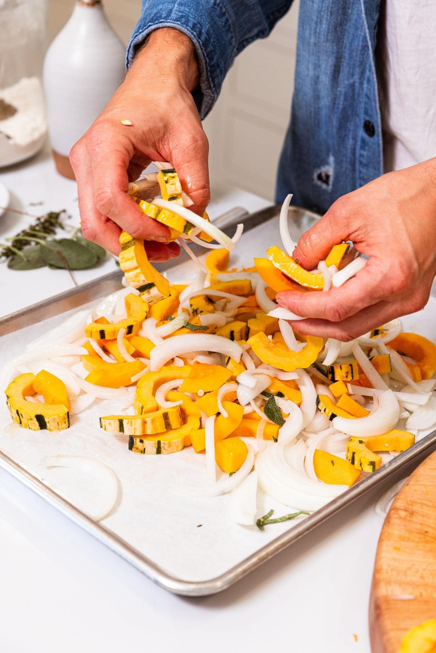 Two hands rubbing olive oil into pieces of squash and onion on parchment-lined baking sheet on white countertop.