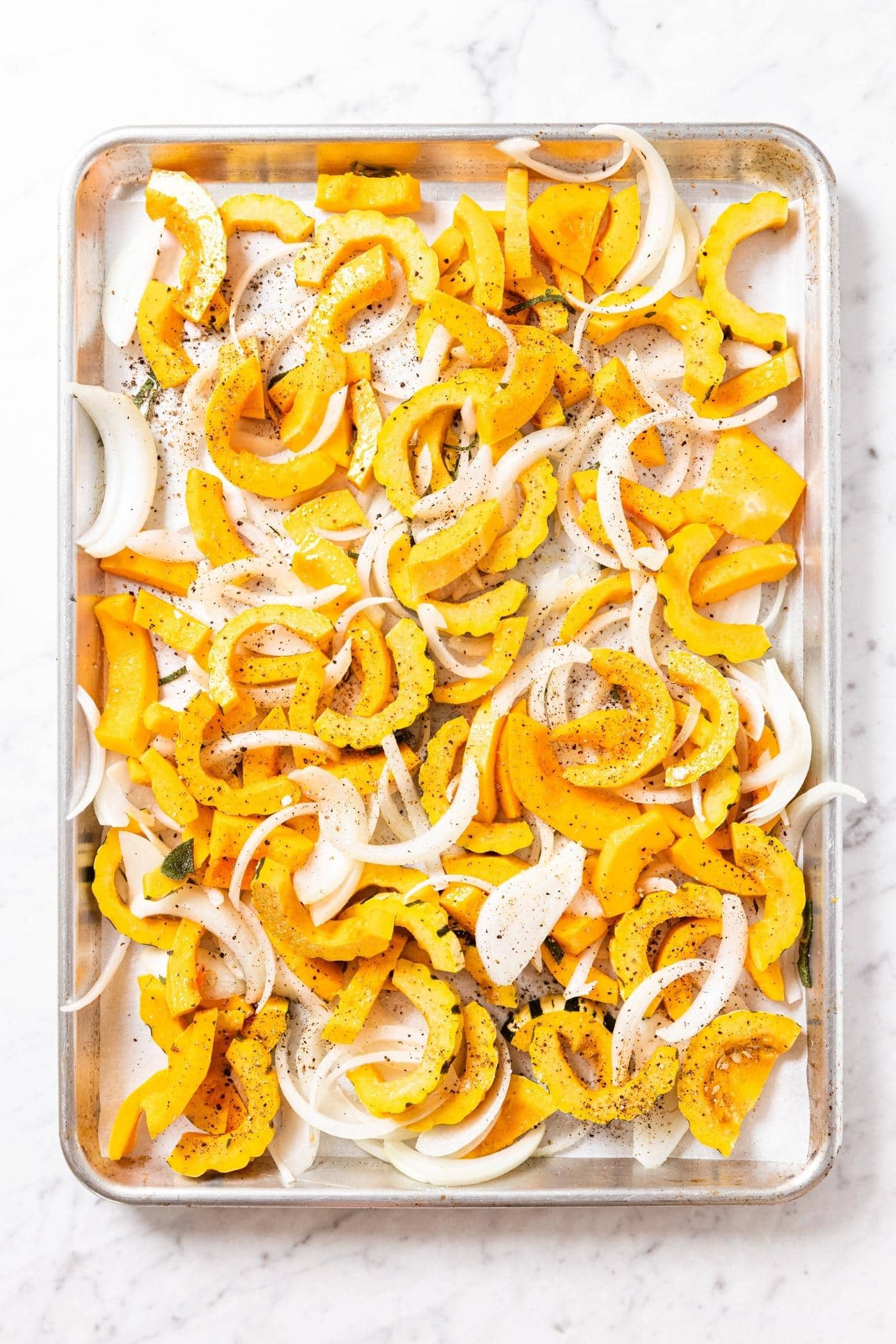 Metal baking sheet sitting on marble surface with uncooked squash and onion slices drizzled with olive oil and herbs.