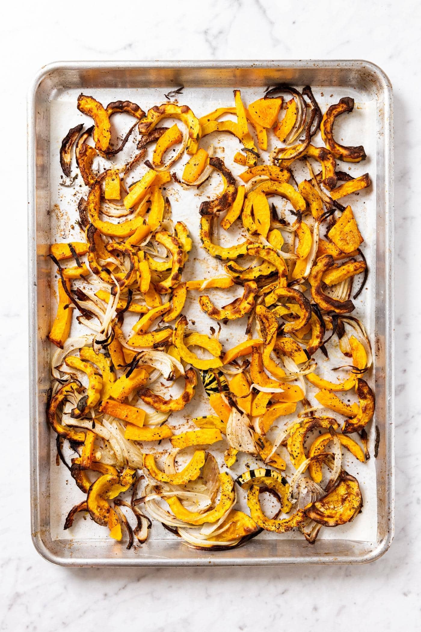 Metal pan filled with roasted pieces of squash and onion showing the slightly browned bits all on marble surface.