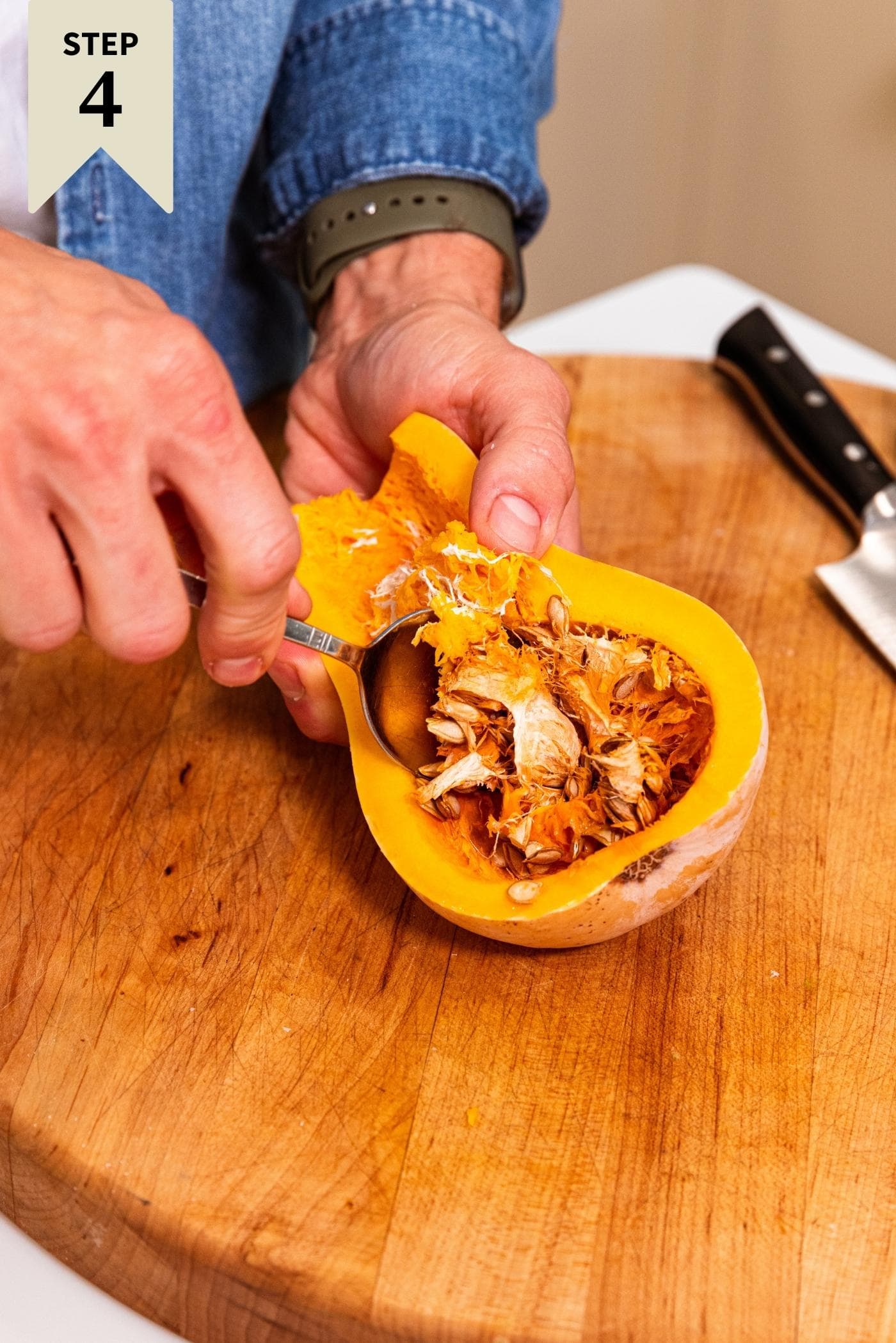 Hand scooping out seeds from interior of butternut squash with wood cutting board underneath.